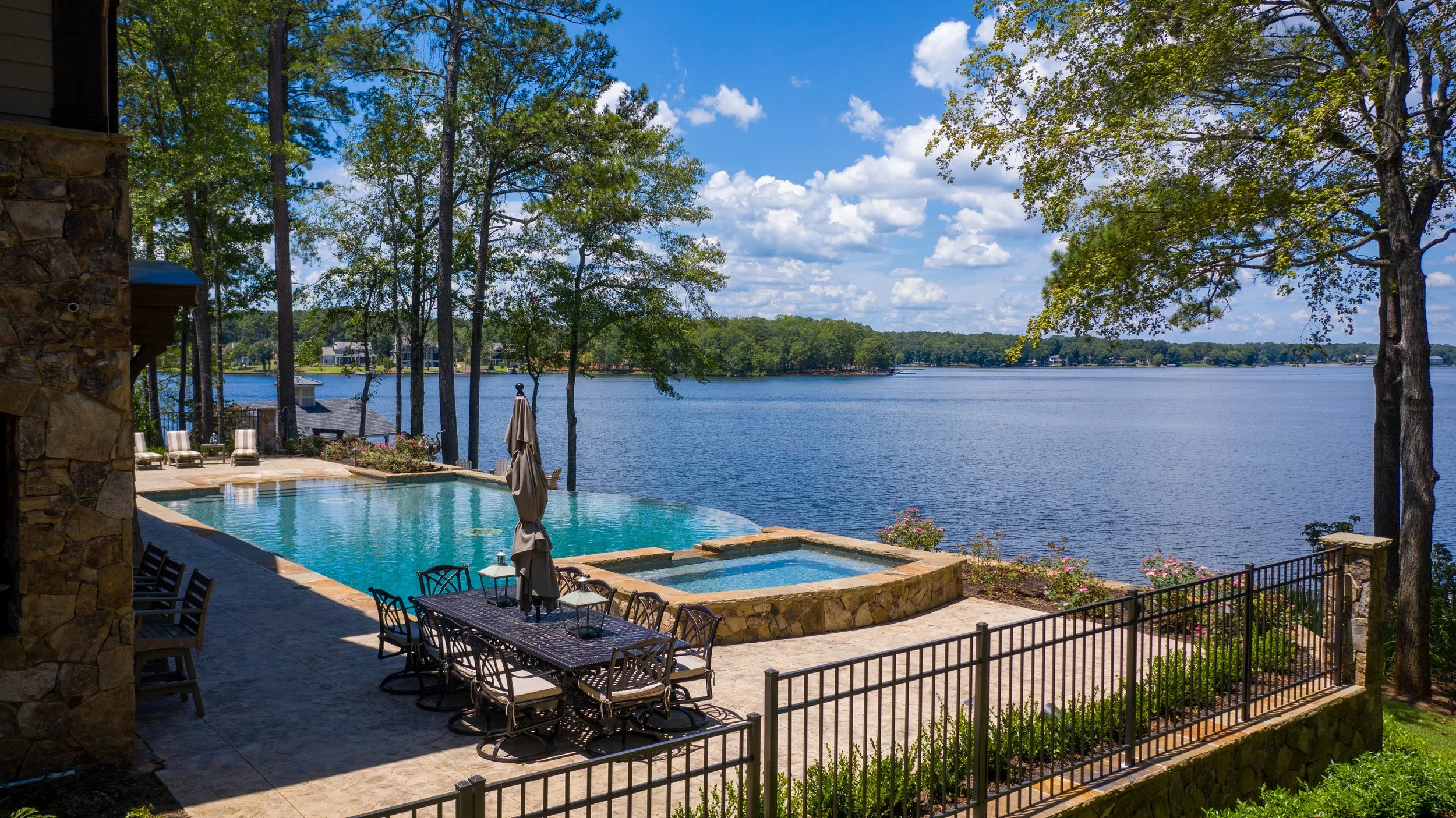 View of a backyard with a swimming pool, hot tub, outdoor dining table, and chairs, overlooking a lake with trees and houses in the background under a blue sky with clouds.