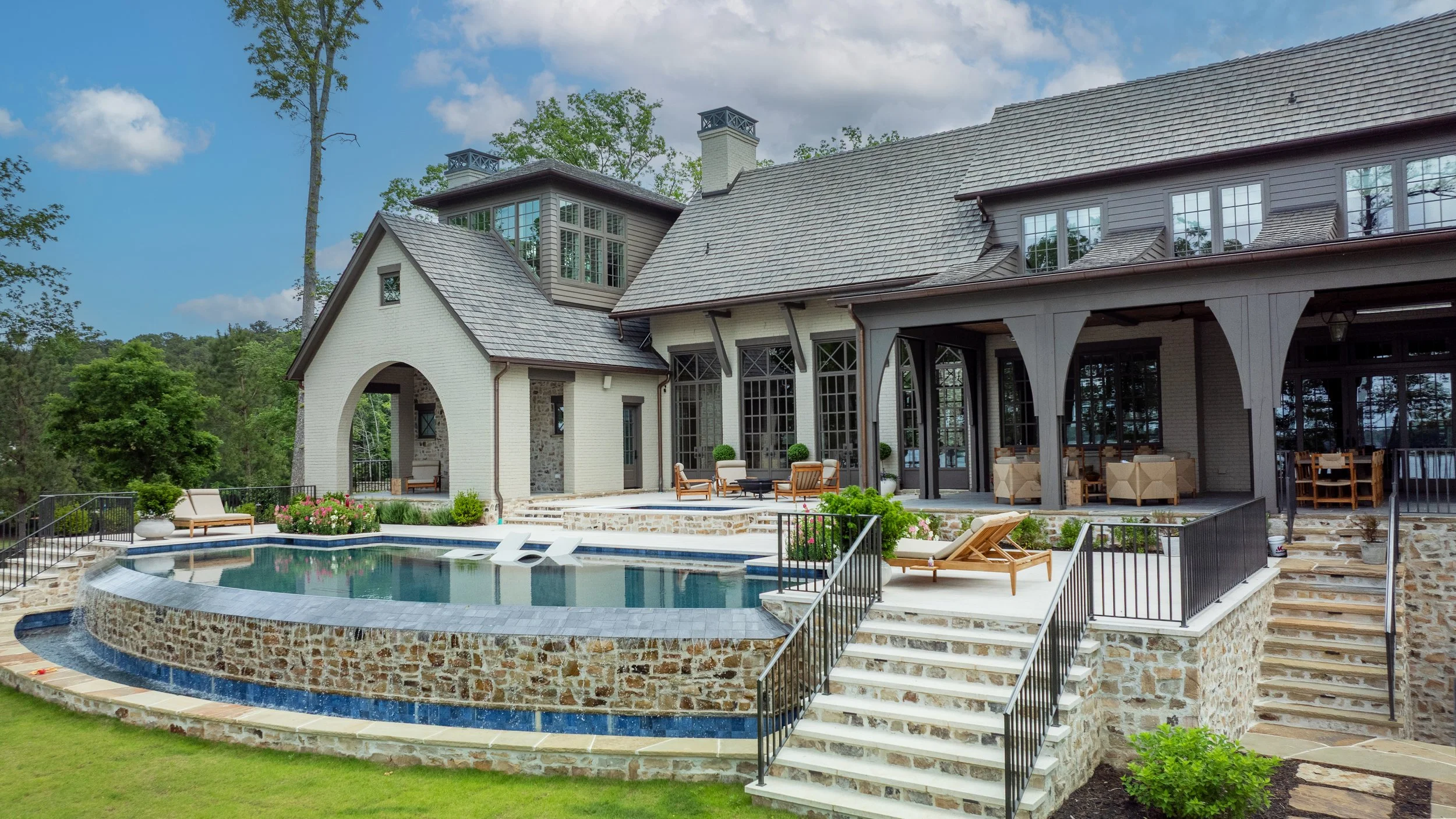 Large house with multiple windows, outdoor patio, and a swimming pool surrounded by stone and metal railings, with outdoor furniture and a lush green background.