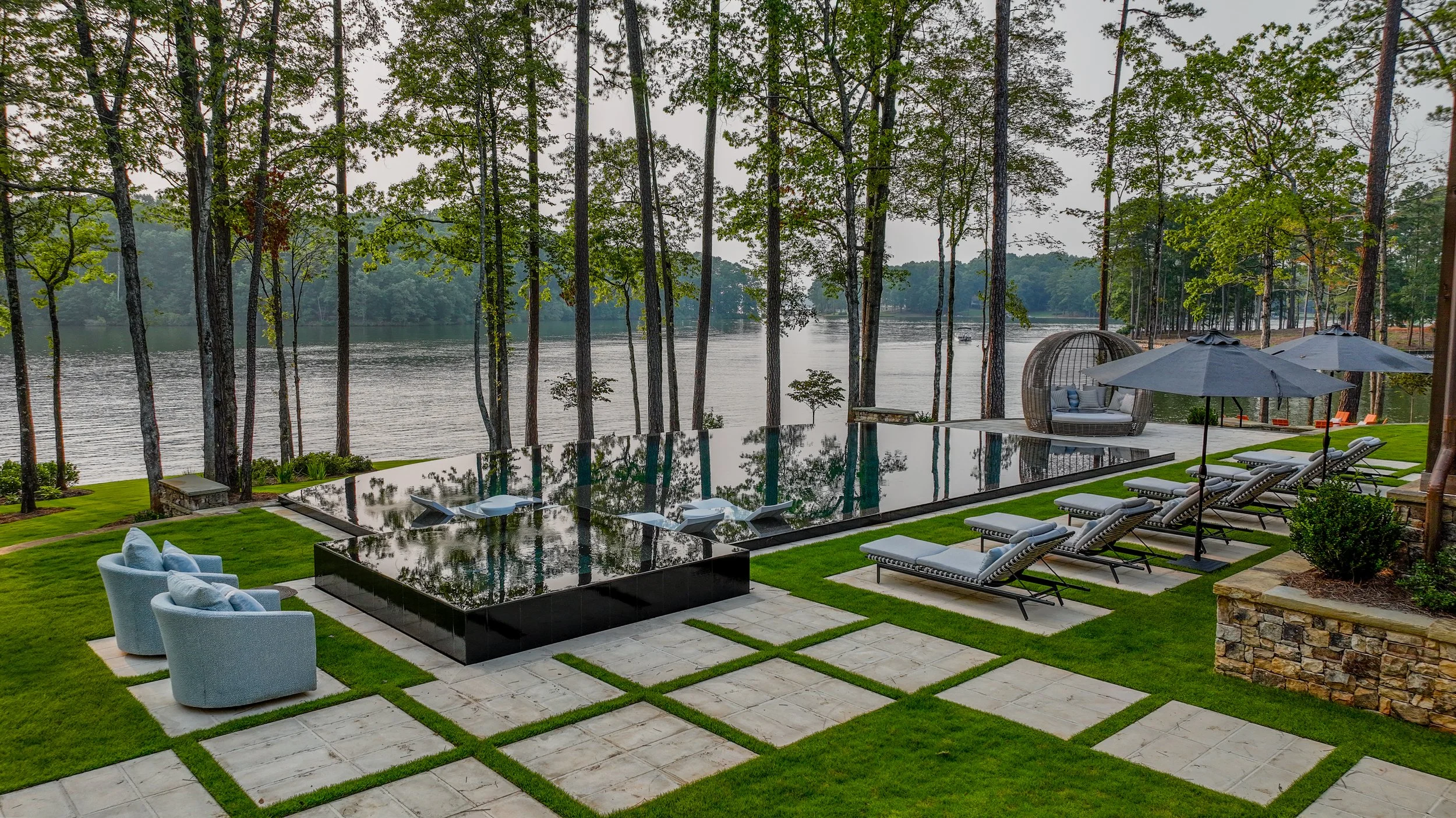 A luxurious outdoor patio with a reflective black pool, surrounded by green grass and stone tiles. Lounge chairs, umbrellas, and outdoor seating are visible, overlooking a river with tall trees in the background.