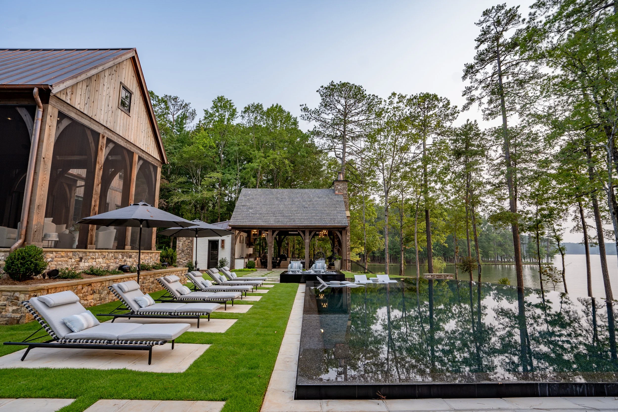 Luxury lakeside backyard with pool, lounge chairs, umbrellas, and a wooden house and pavilion, surrounded by trees and water.