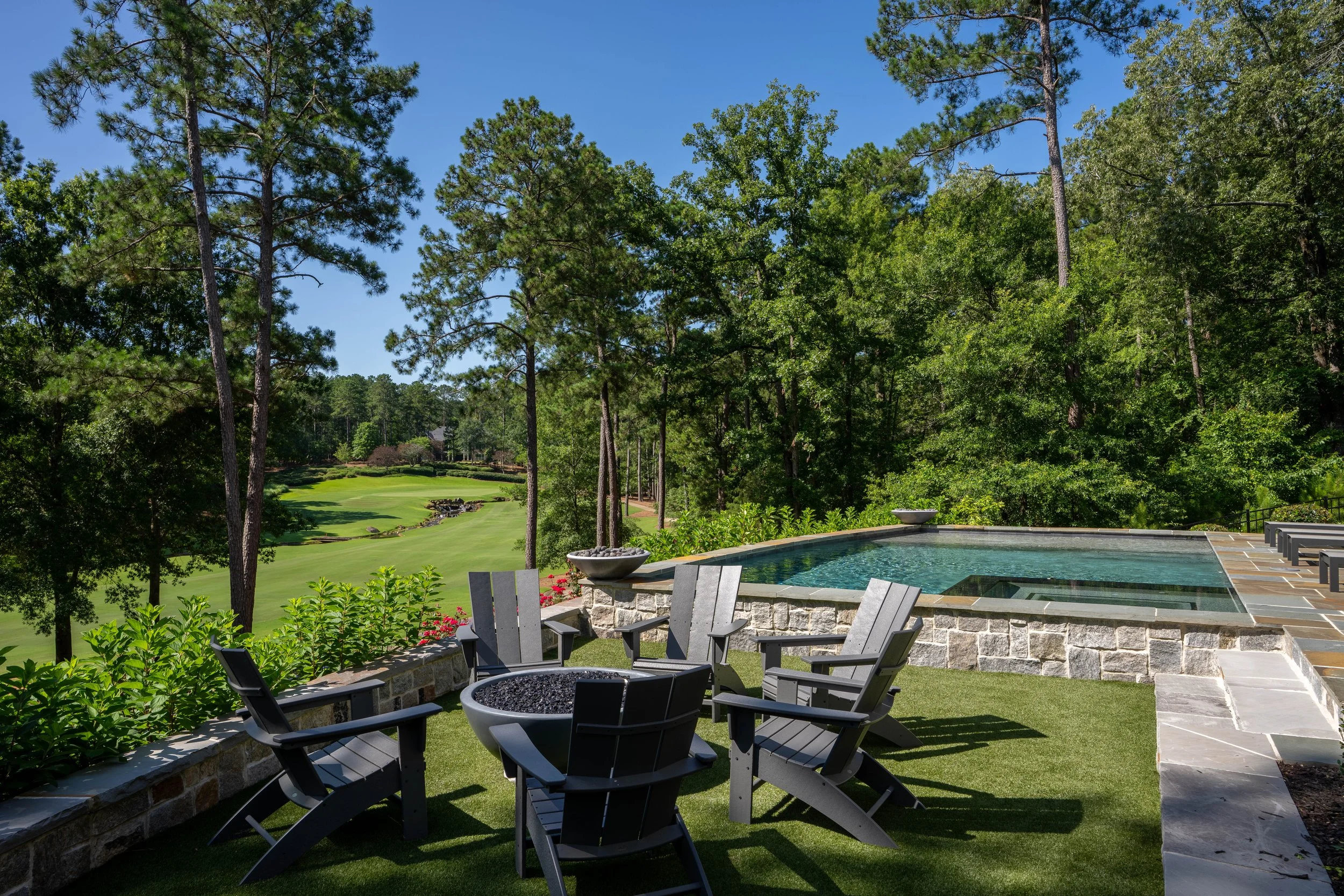 Luxury outdoor patio with a hot tub, six chairs, and a fire pit, overlooking a lush green golf course surrounded by tall trees on a clear, sunny day.