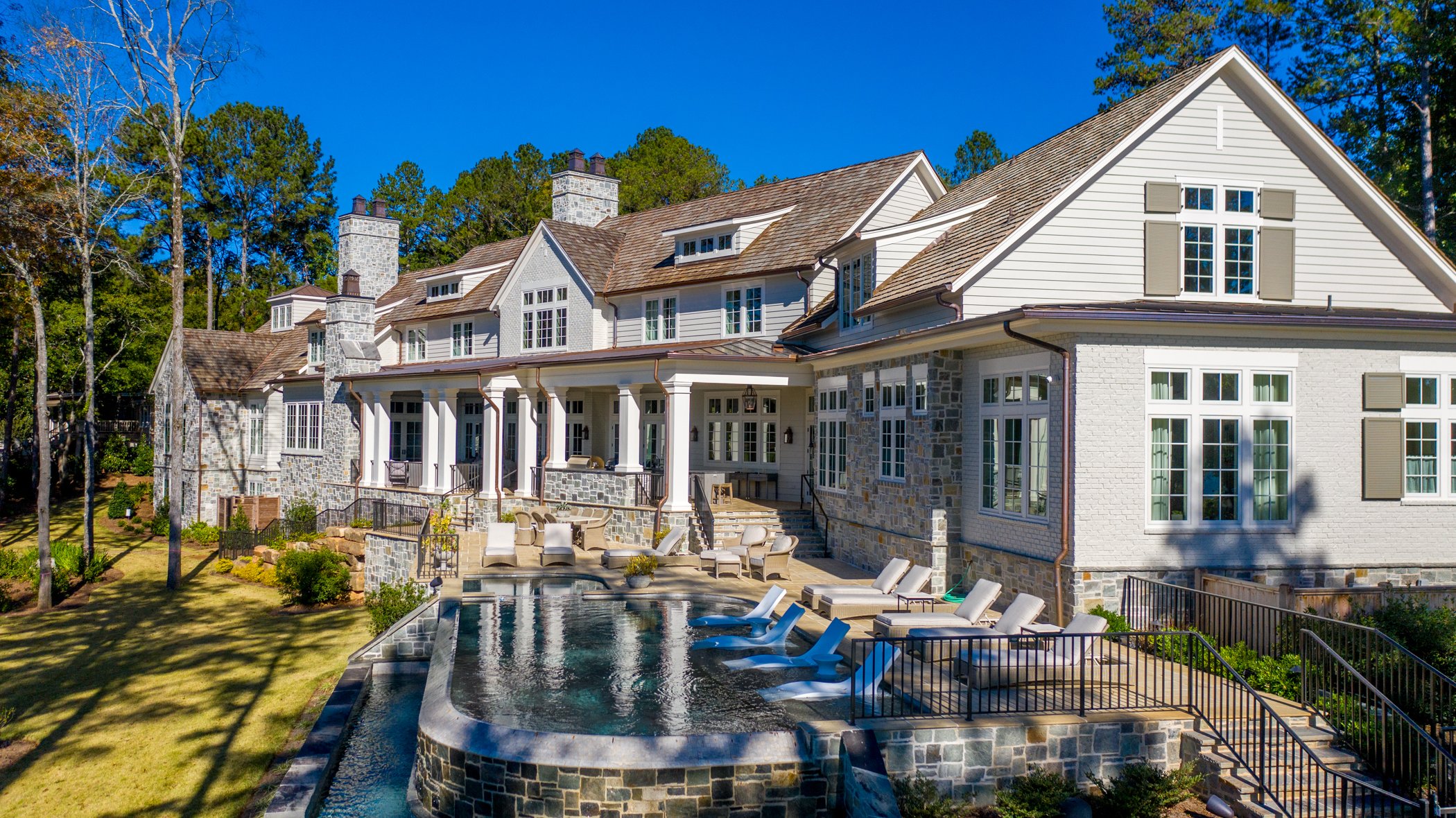 Large luxury house with white walls, multiple windows, and stone accents, featuring a swimming pool in the backyard with lounge chairs and outdoor seating under a clear blue sky.