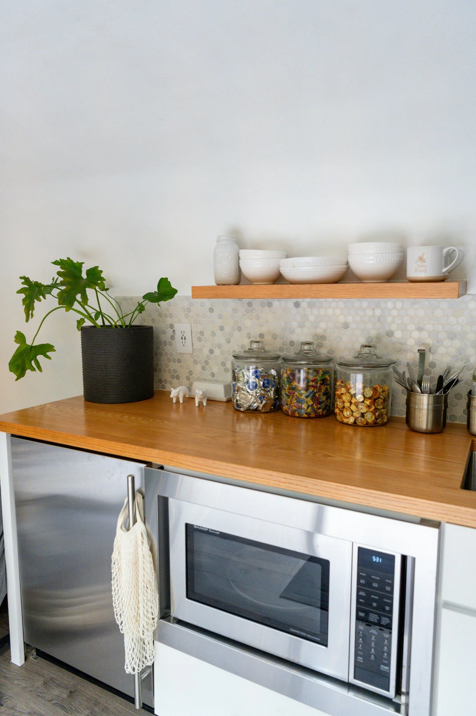 Kitchen counter with a black potted plant, three ceramic white bowls and a mug on a floating wooden shelf, three glass jars filled with candies or chocolates, a stainless steel utensil holder with spoons and forks, a microwave, and a white cabinet below.