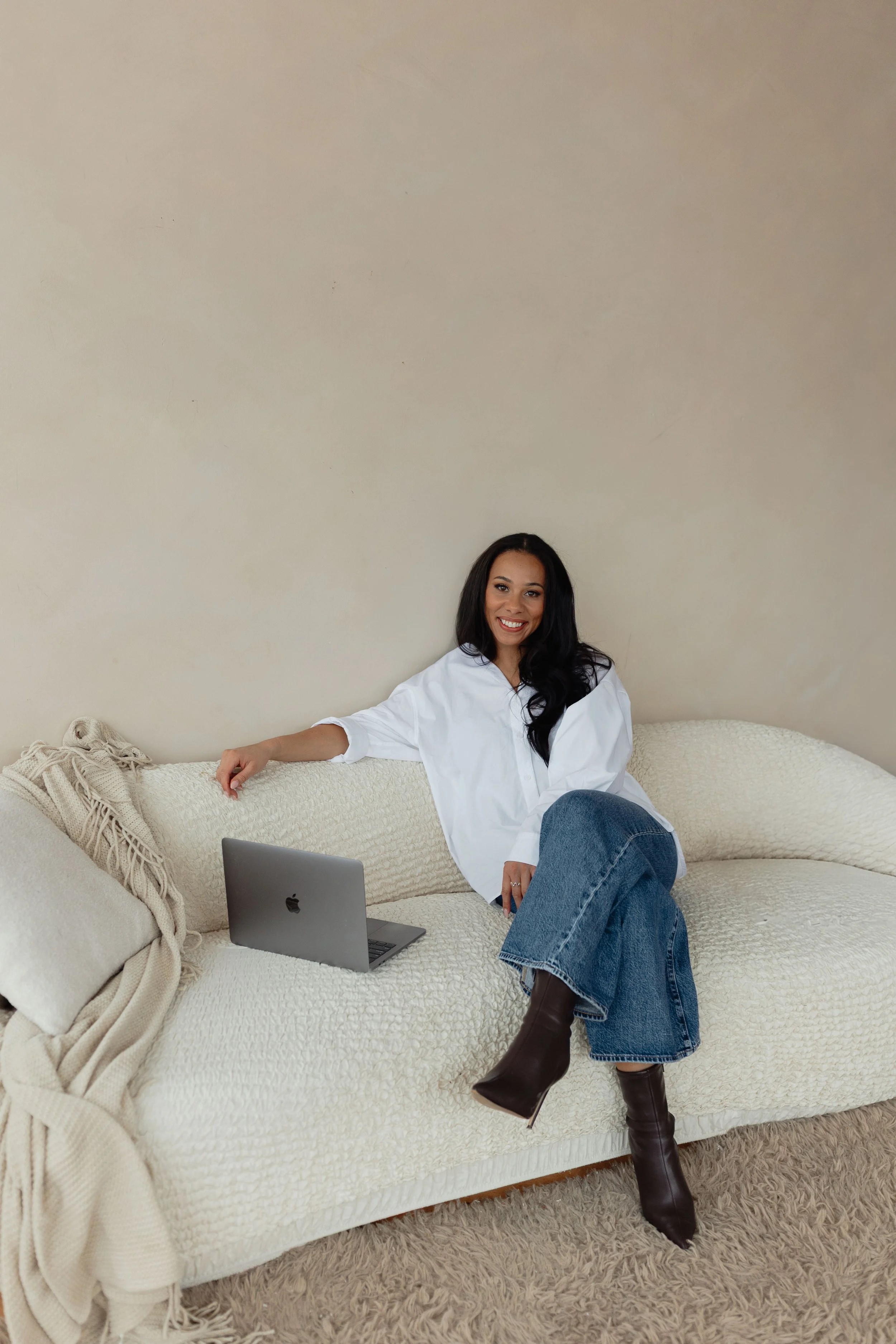 A woman sitting on a cream-colored sofa with a laptop, smiling at the camera, in a neutral-colored room.