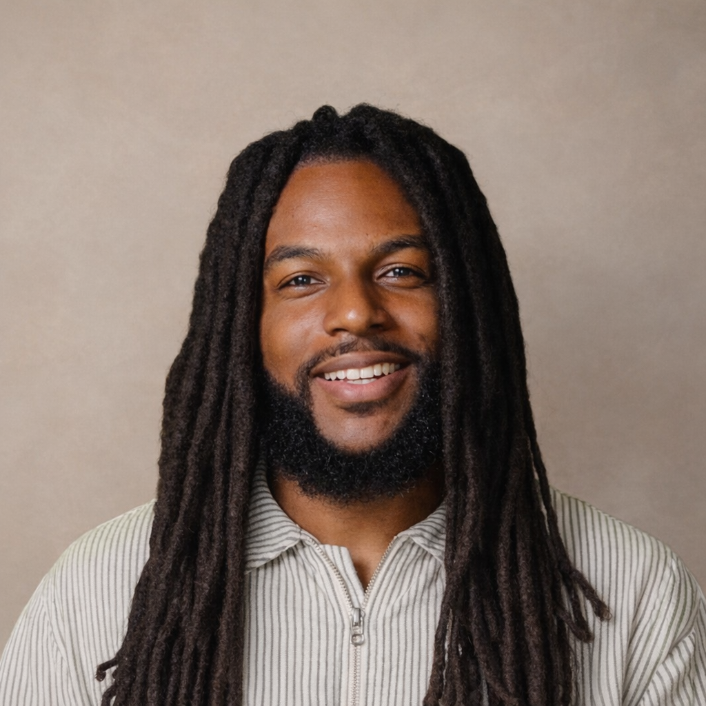 Portrait of a man with long dreadlocks, a beard, smiling, wearing a light-colored, striped zip-up shirt, against a plain neutral background.
