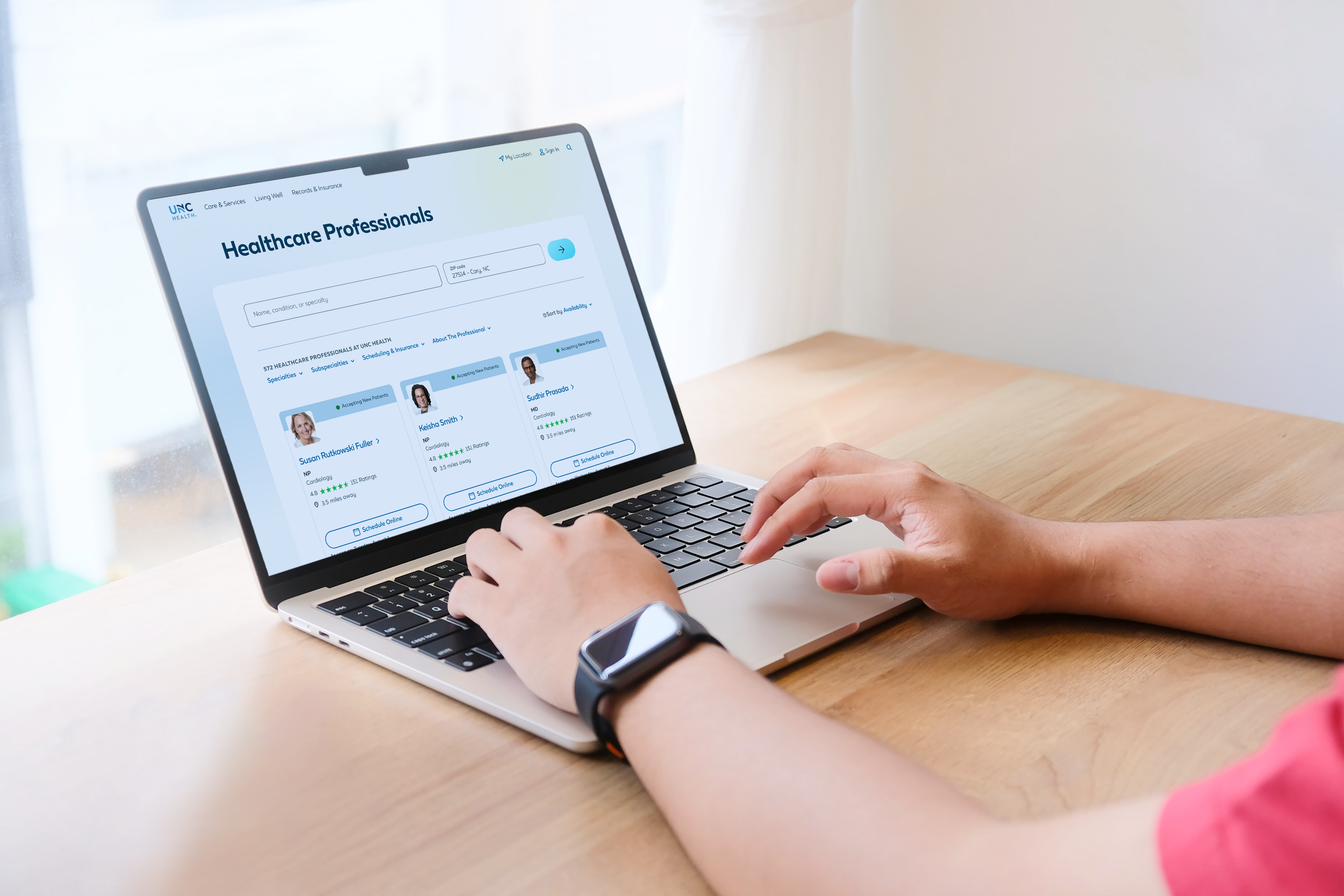 Person wearing a smartwatch using a laptop on a wooden table, viewing a healthcare professional directory online.