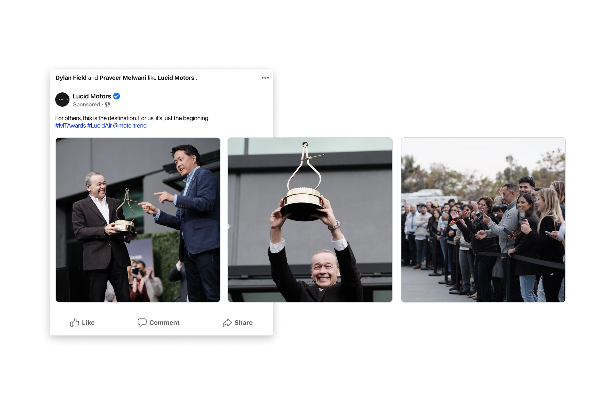 A man in a suit holding a trophy while another man in a suit is smiling and pointing at him, with a group of people in line watching and taking photos in the background.
