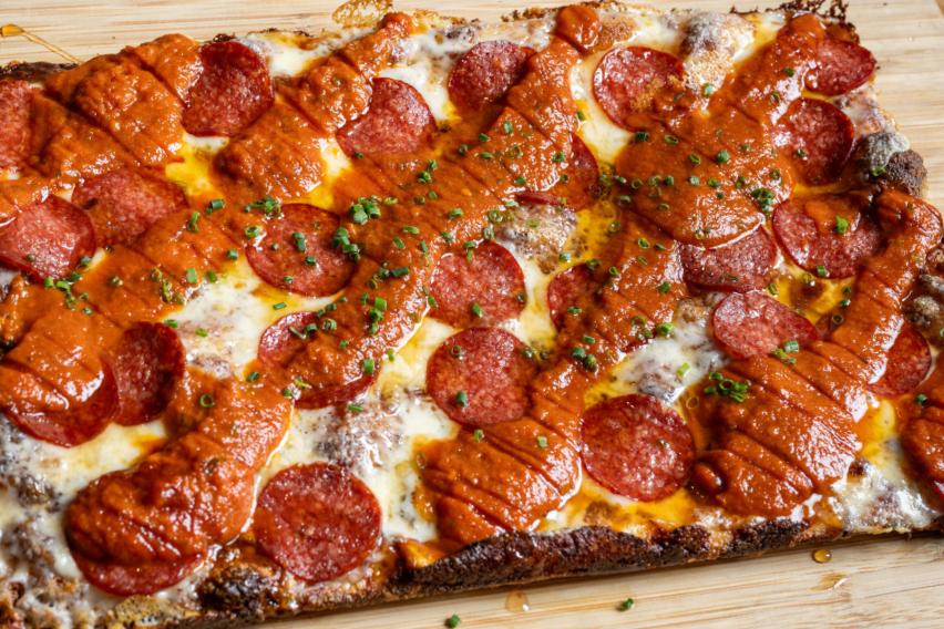 Close-up of a 9MILE Detroit Style Square Pizza pepperoni and sausage with melted cheese and chopped green herbs on a wooden surface.