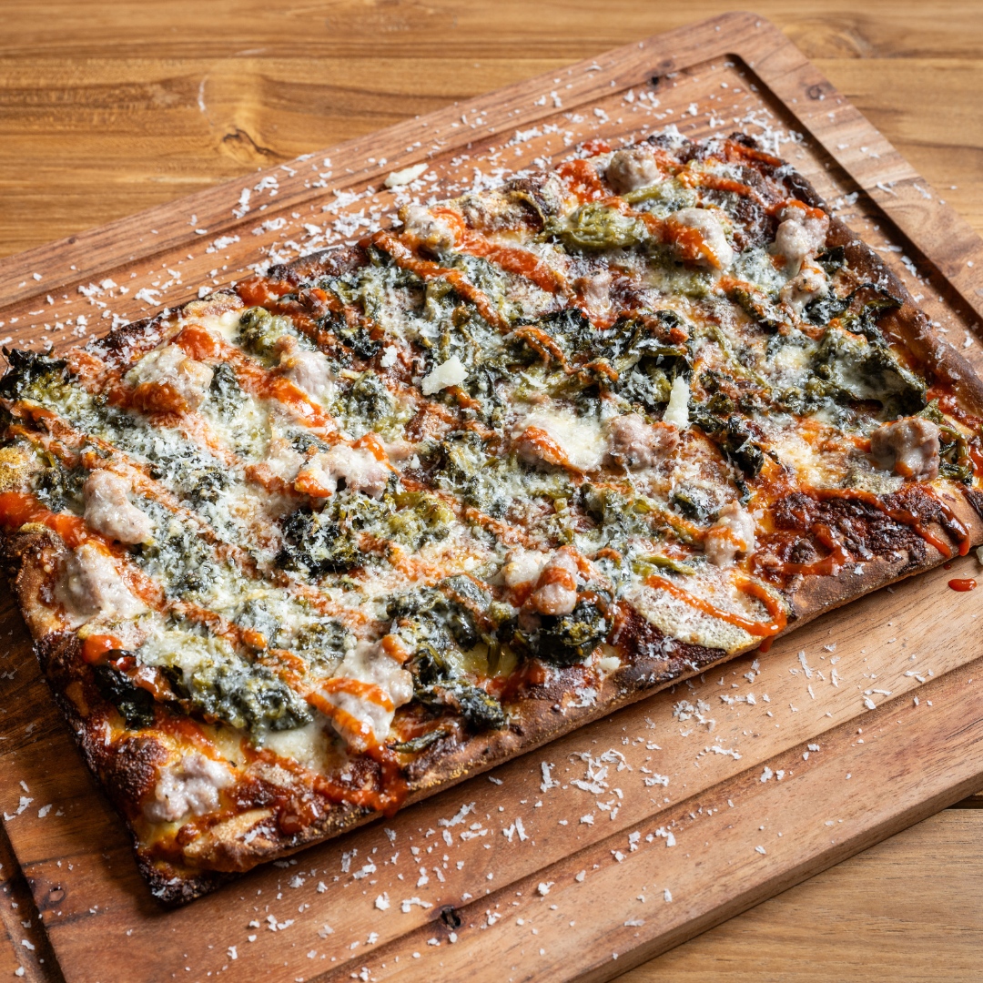 A rectangular 9MILE Detroit Style Pizza topped with cheese, green peppers, sausage, and drizzled with hot sauce on a wooden pizza board.
