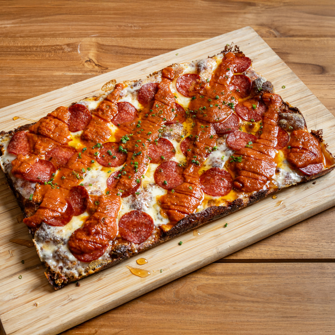 9MILE Detroit Style Square Pizza topped with pepperoni slices, sausage, melted cheese, and chopped green herbs on a wooden cutting board.
