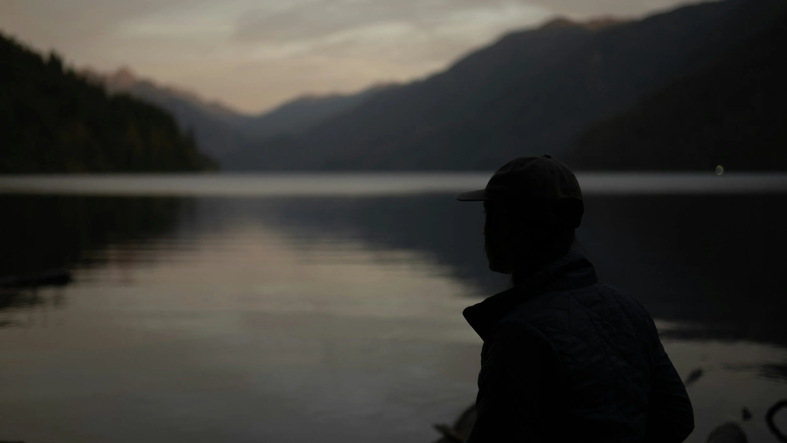 Person wearing a cap and jacket sitting by a calm body of water at dusk, with mountains in the background.