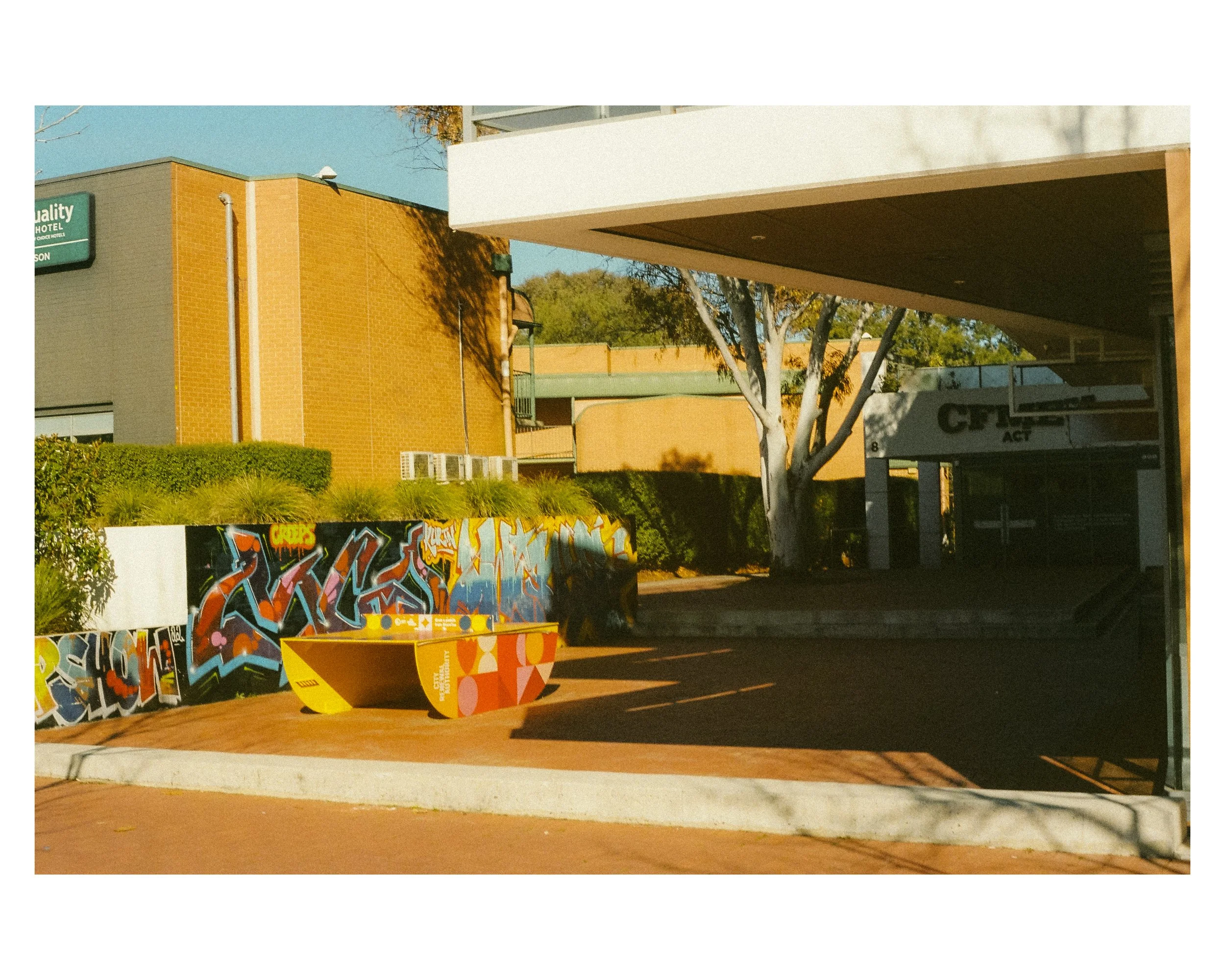 An outdoor urban scene with a colorful graffiti-covered wall, a yellow art installation, a tree, and parts of buildings including a hotel and a parking garage, during daylight with shadows cast on the ground.