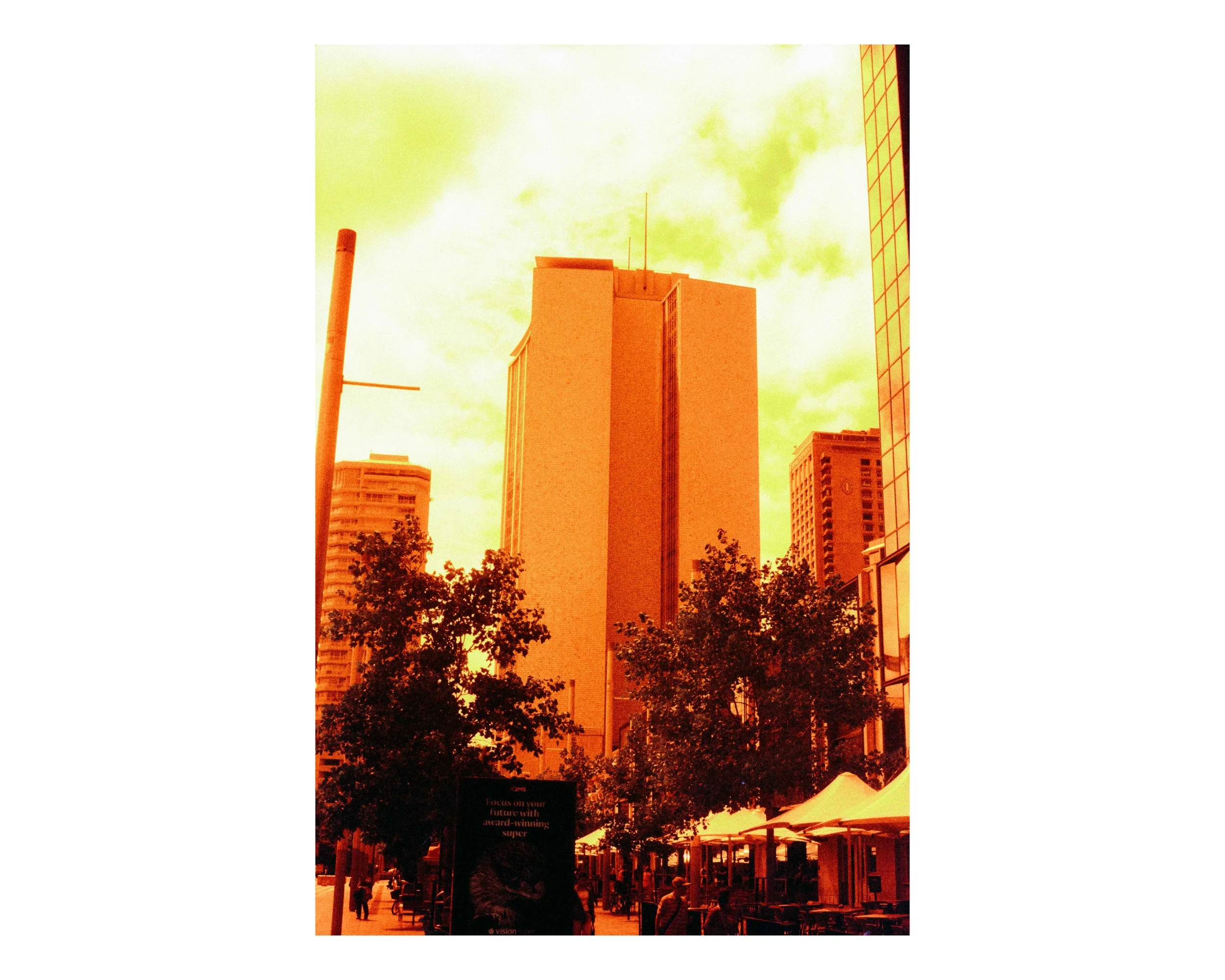 A city street with tall buildings, trees, and outdoor cafes with umbrellas, under a yellowish sky.
