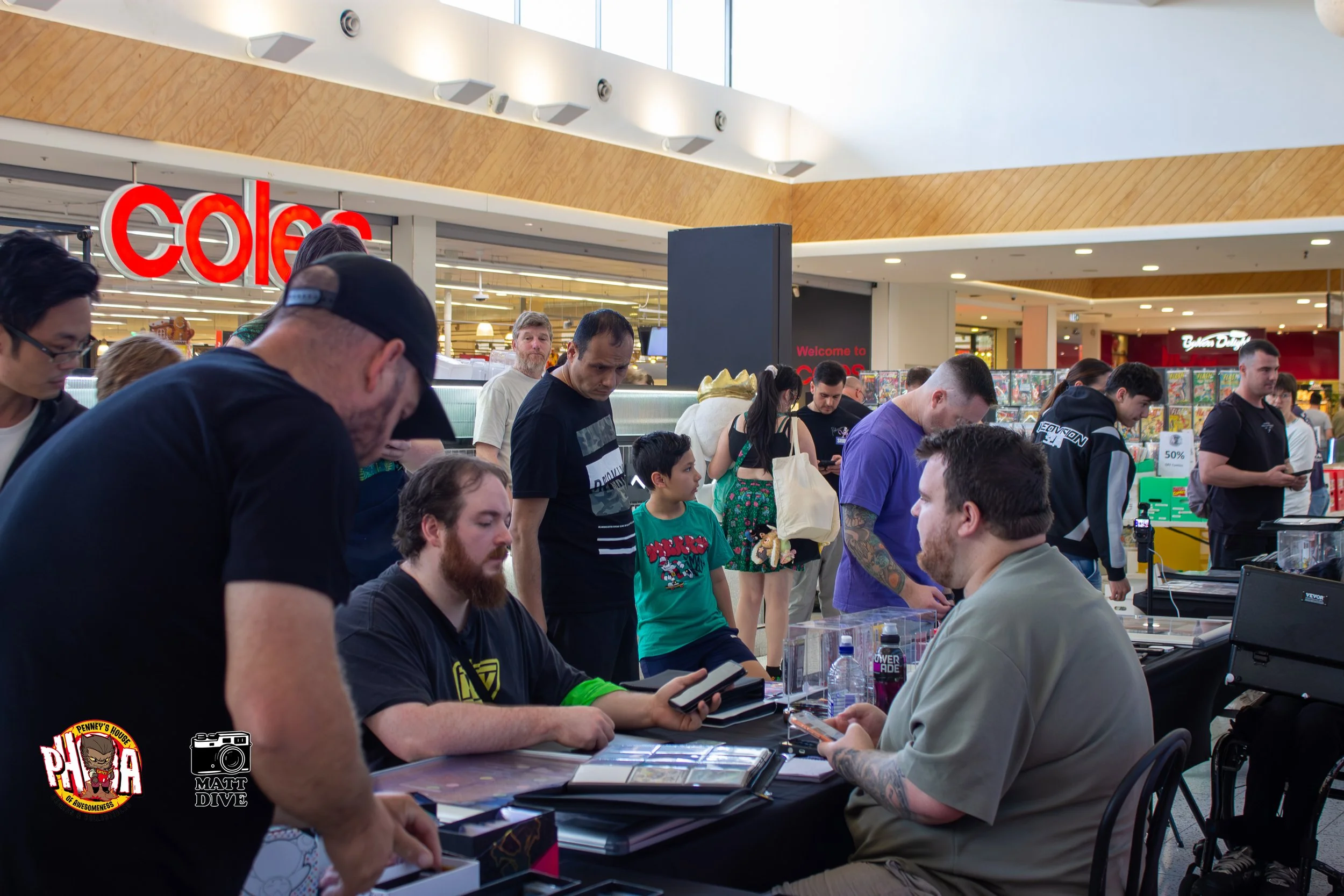 People in line at a card game event inside a shopping mall, with a vendor selling cards and others browsing and managing the event.
