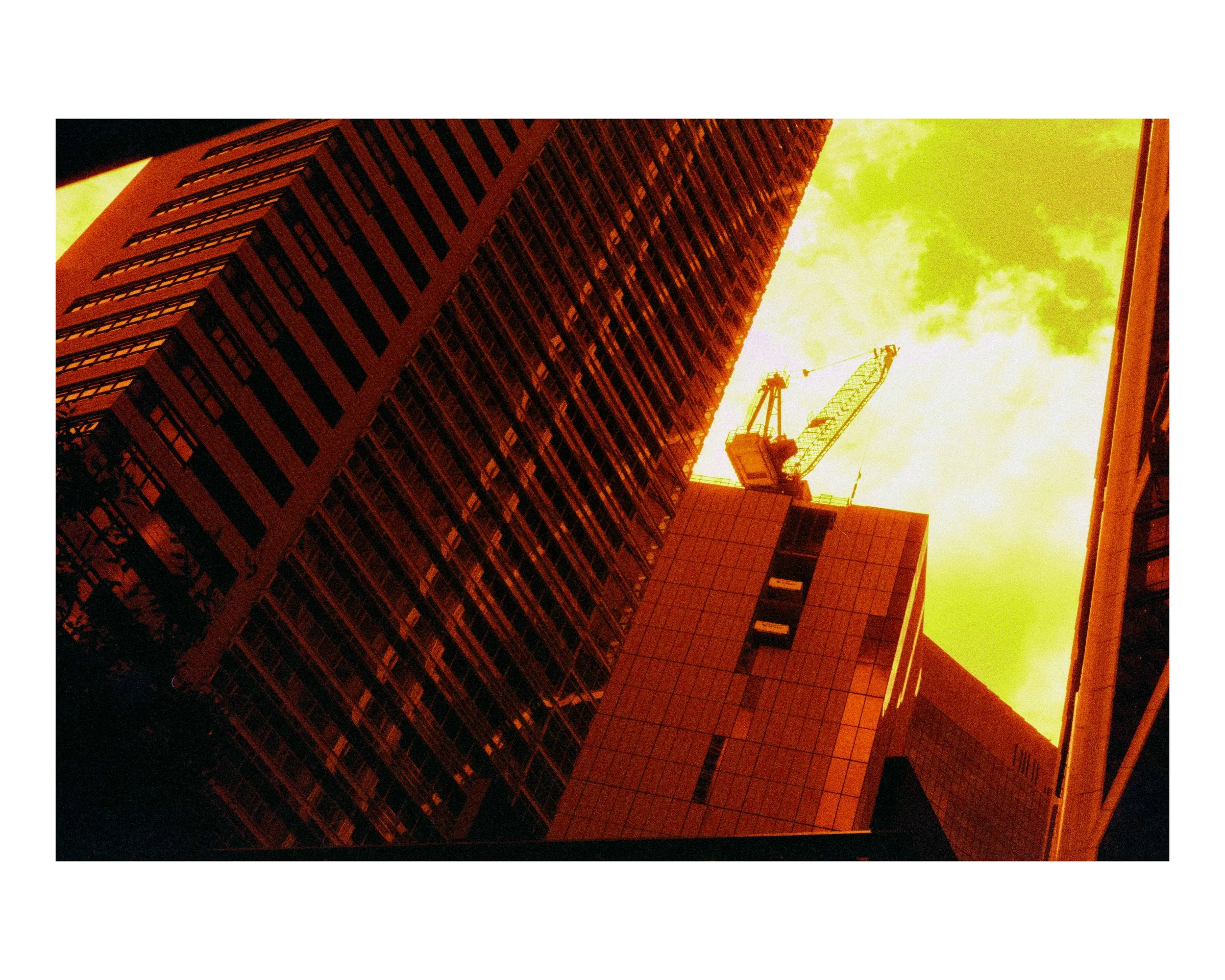 Looking up at skyscrapers and a crane against an orange and green sky.