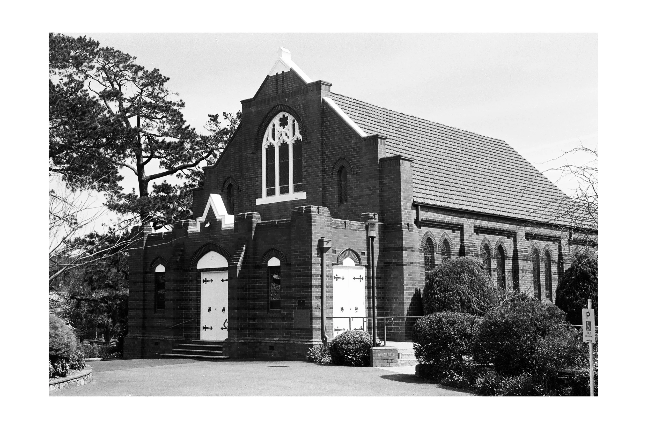 Black and white photo of a brick church with arched windows, a steep roof, and a small set of stairs leading to double doors.