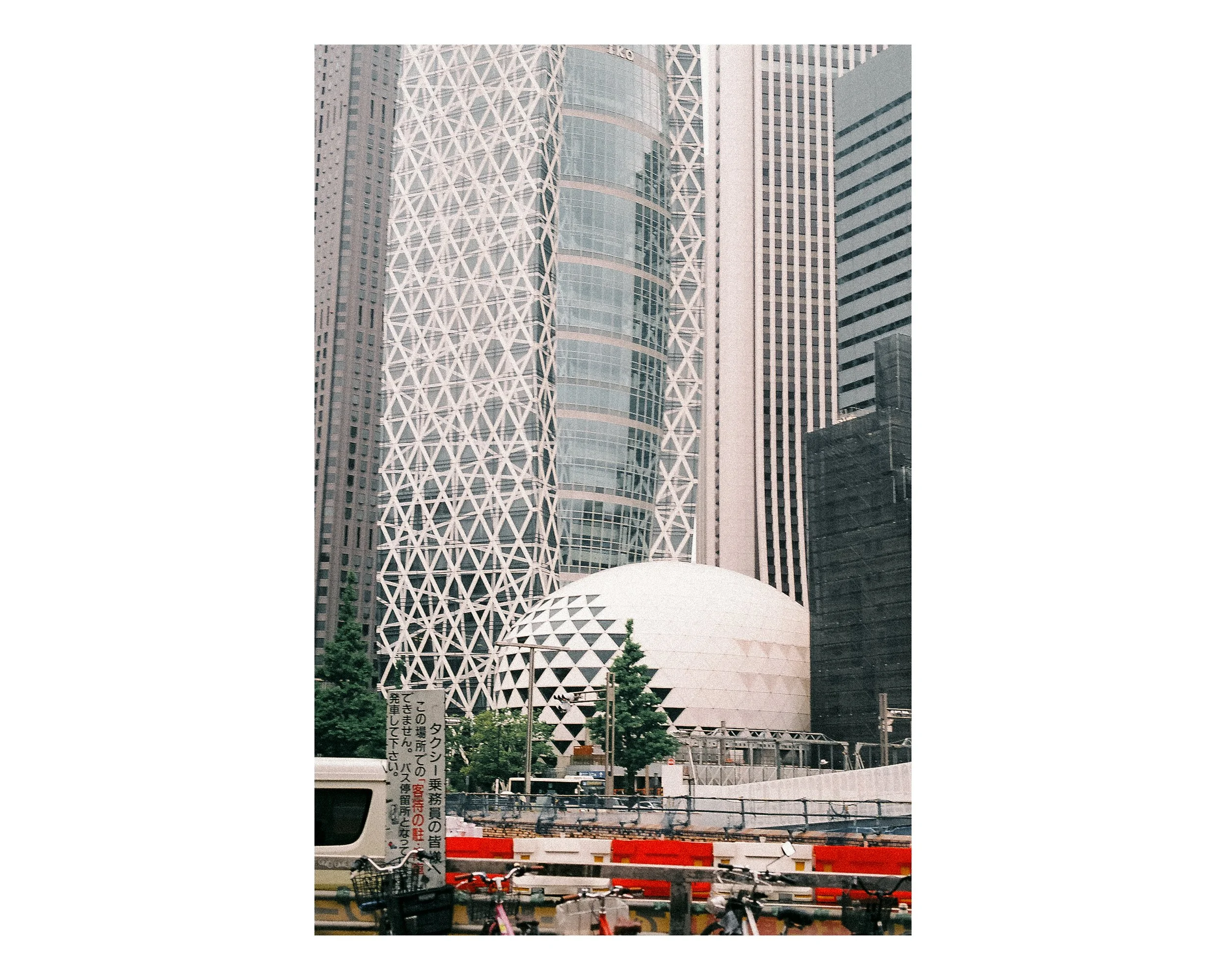 Tall modern skyscrapers in an urban area with a white geometric dome structure at their base, trees, construction barriers, bicycles, and a sign with Japanese characters.
