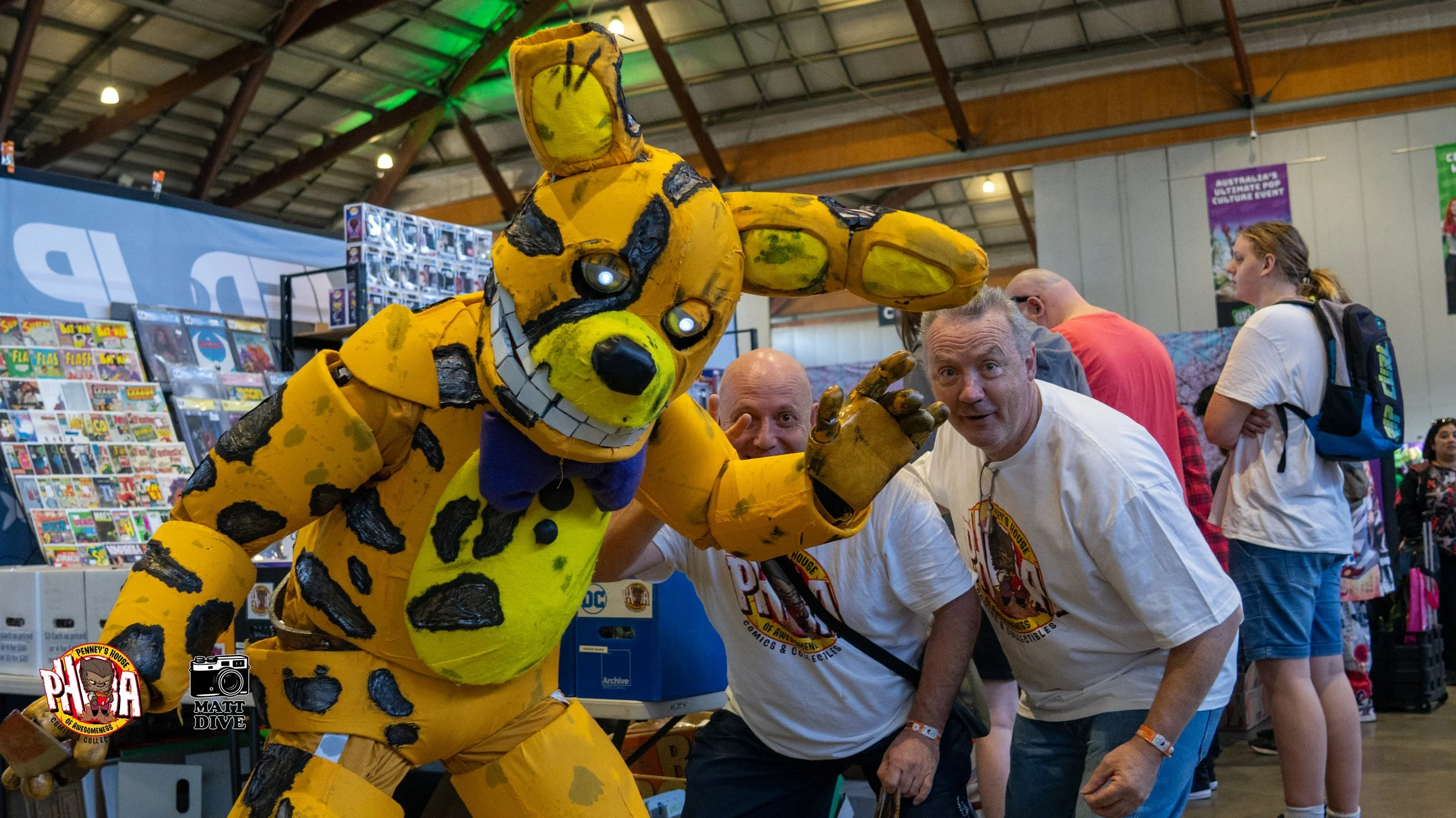 Two men with grey hair and white t-shirts smiling and posing with a brightly colored, large, custom robotic mascot resembling a cartoonish dog at a comic convention or event. The mascot is yellow with black spots, a big smile, and is pointing towards