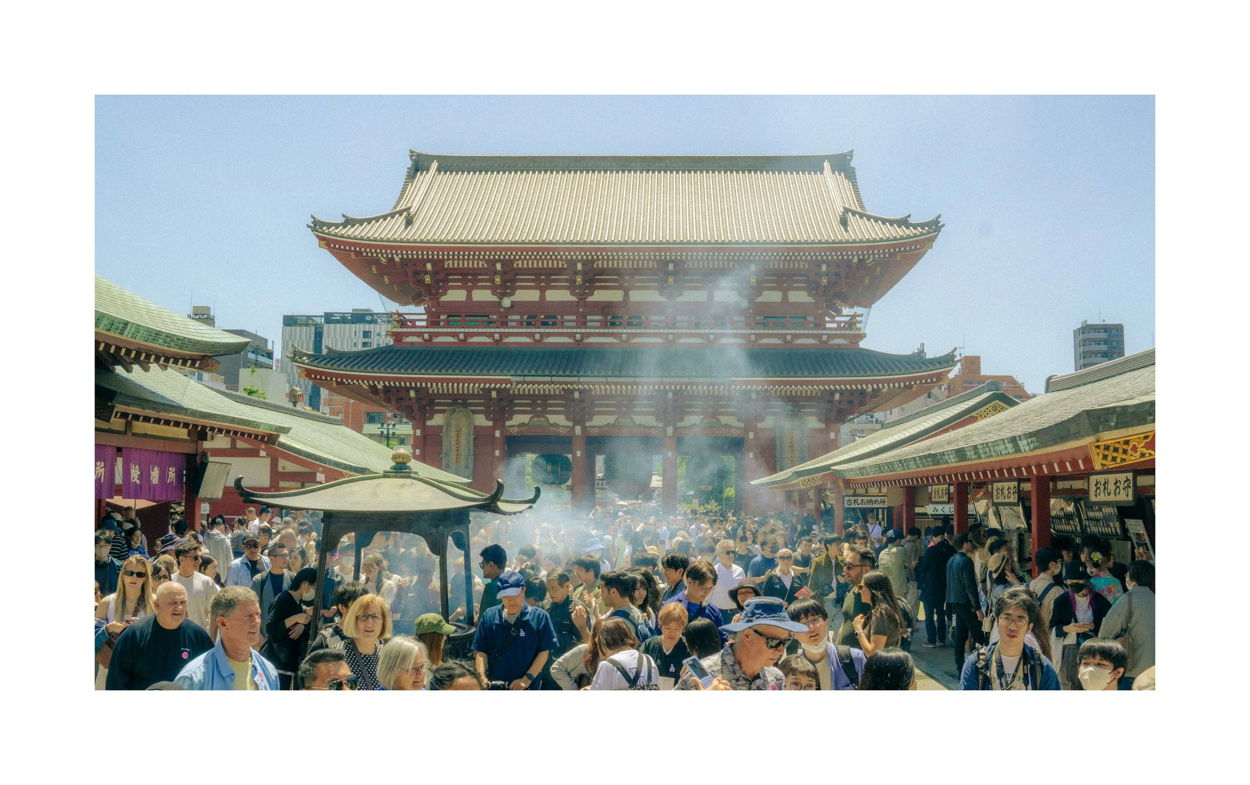 A crowded outdoor scene at a traditional Japanese temple, with many tourists and visitors. The temple has a large, ornate pagoda-style structure with a yellow-tiled roof. Smoke rises from a small incense burner in the foreground, and smaller building