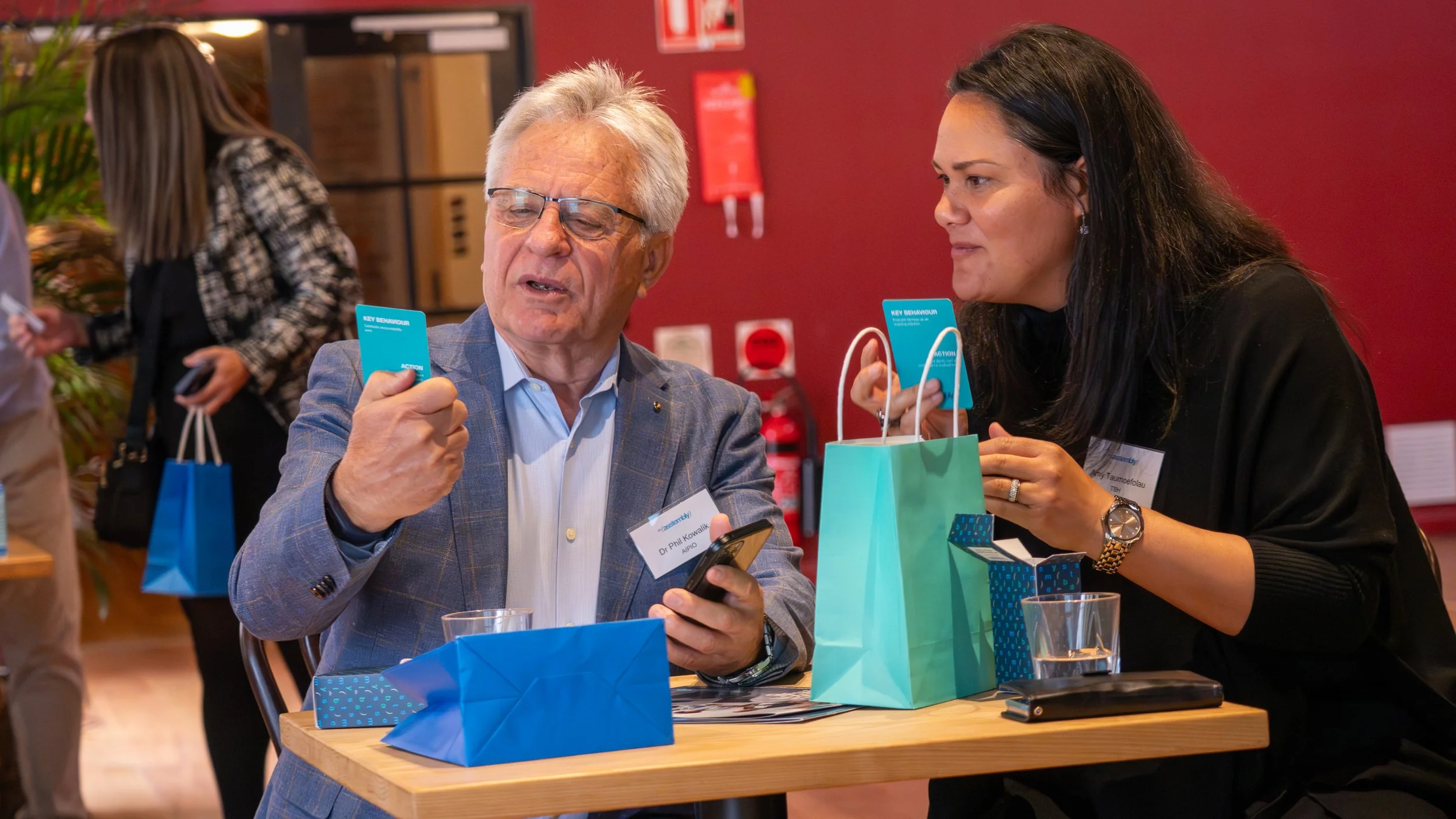 An older man and a woman sit at a table exchanging blue cards. The man is looking at his phone and the woman is holding a blue card. There are gift bags and glasses of water on the table, with other people in the background.