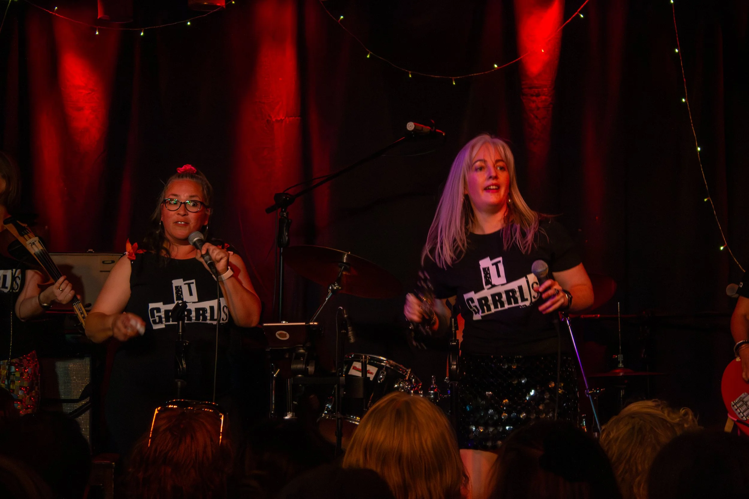 Two women performing singing on stage, holding microphones, with musical instruments and stage lights in the background, and an audience in front.