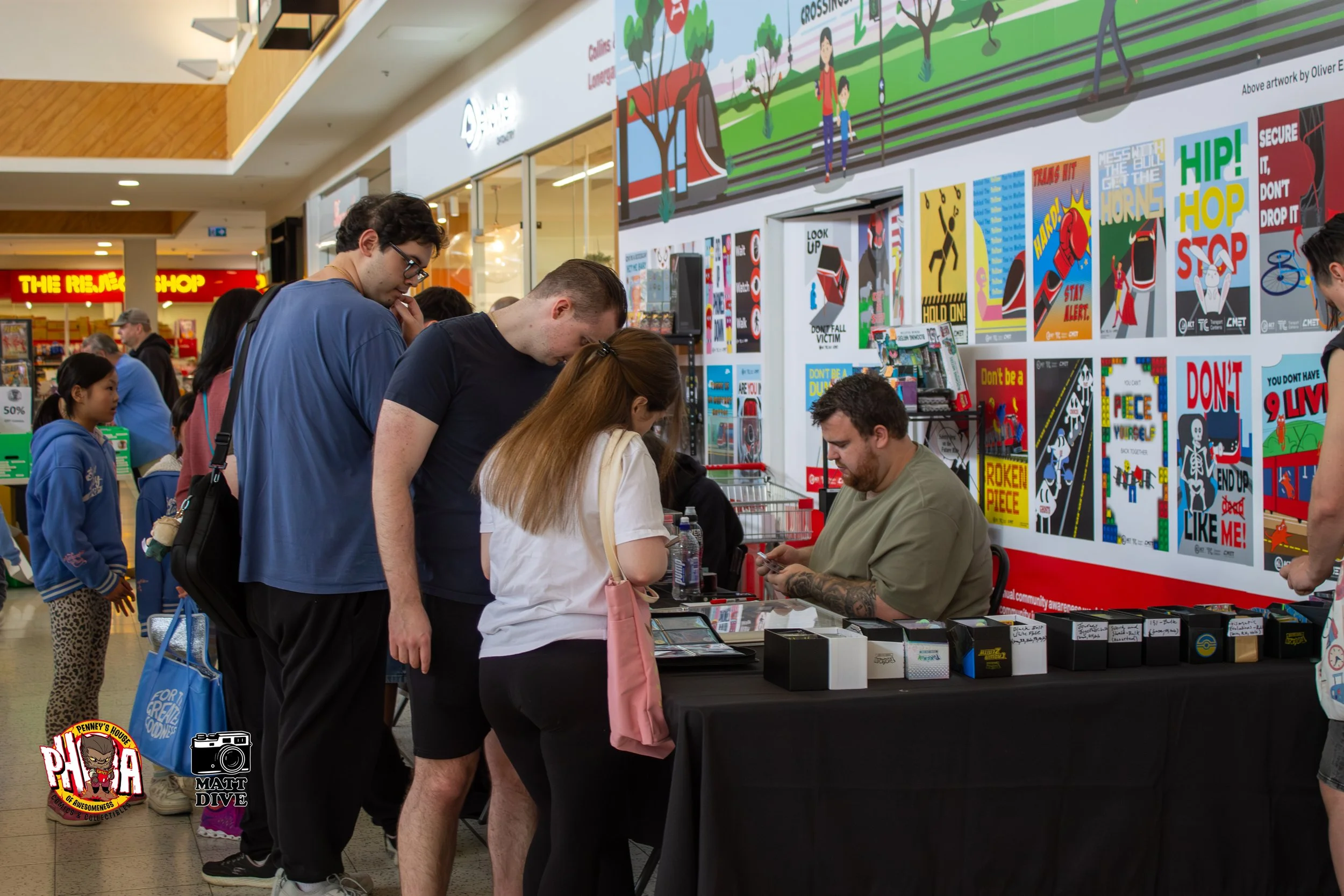 People gathered around a table at a convention or event inside a shopping mall, with posters and artwork displayed on the wall behind them.