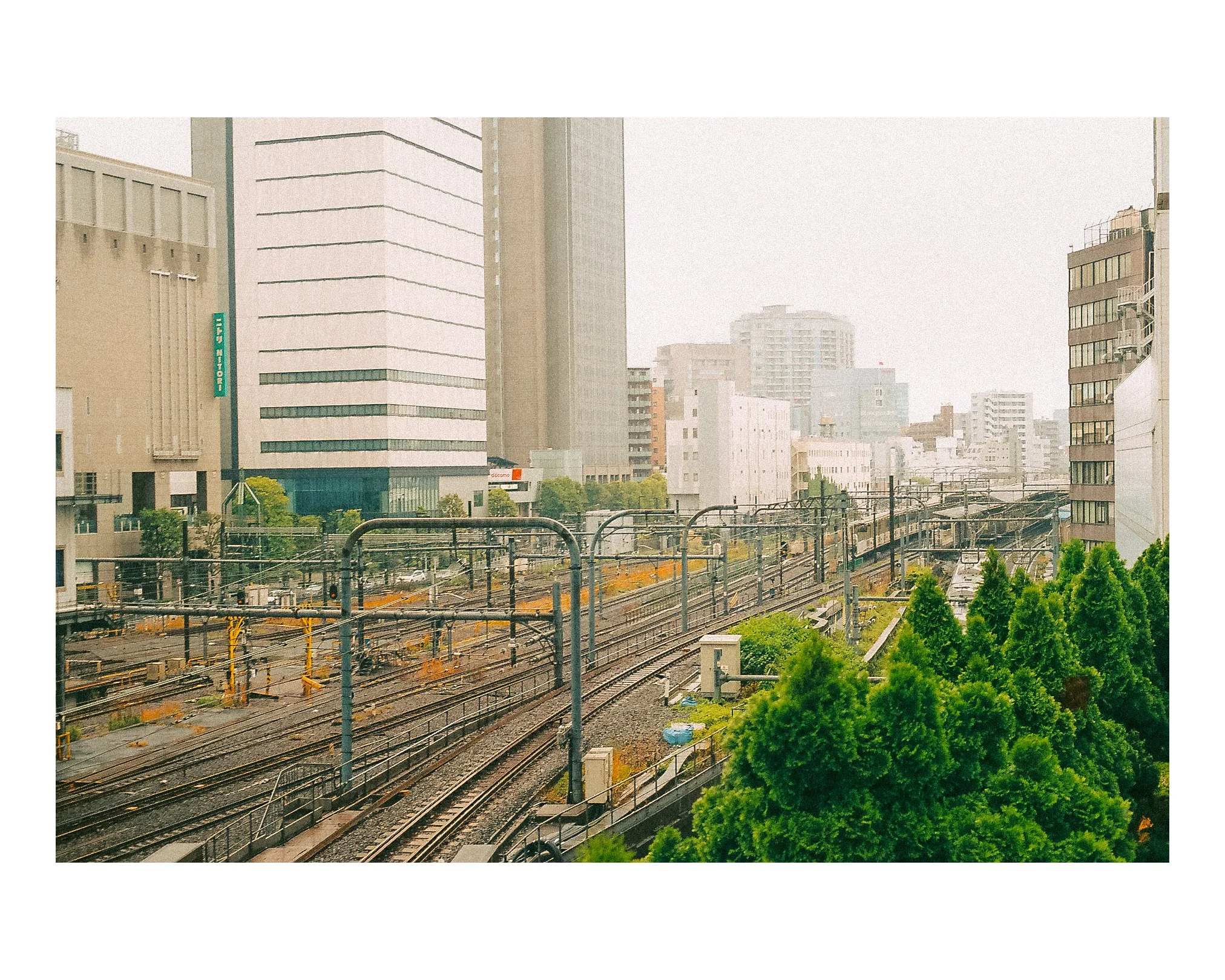 Urban scene with train tracks, overhead electrical wires, and tall buildings in the background on a cloudy day.