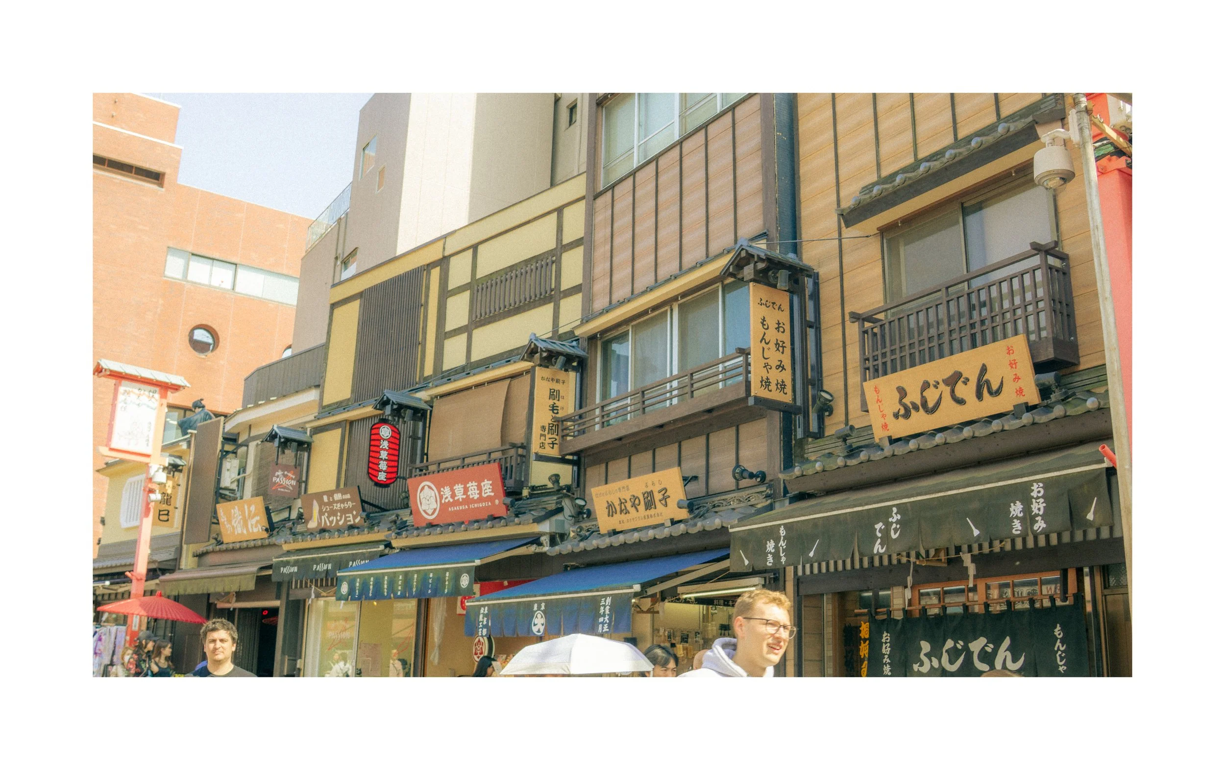 Street view of Japanese storefronts with signs, balconies, and awnings, with people walking, some with umbrellas, and buildings in the background.
