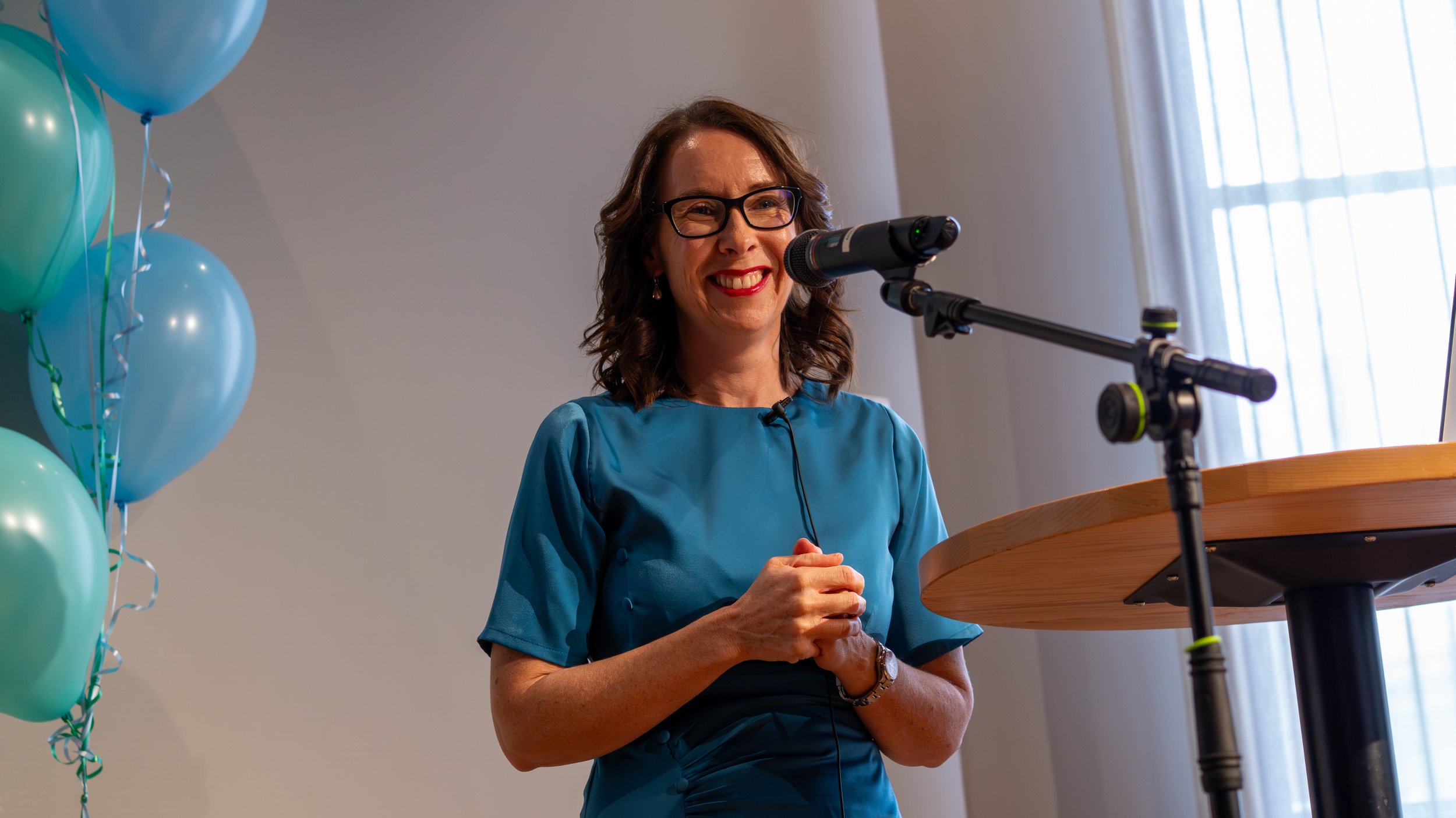 A woman with glasses and dark hair smiling as she stands behind a podium, speaking into a microphone during a presentation or event, with blue and green balloons nearby and sunlight coming through curtains in the background.