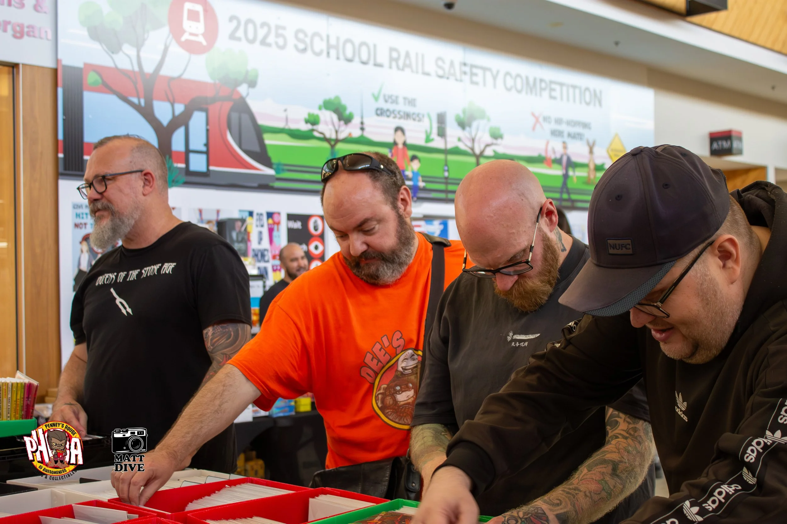 Four men browsing through collectibles at a convention, with a large banner about a 2025 school rail safety competition in the background.