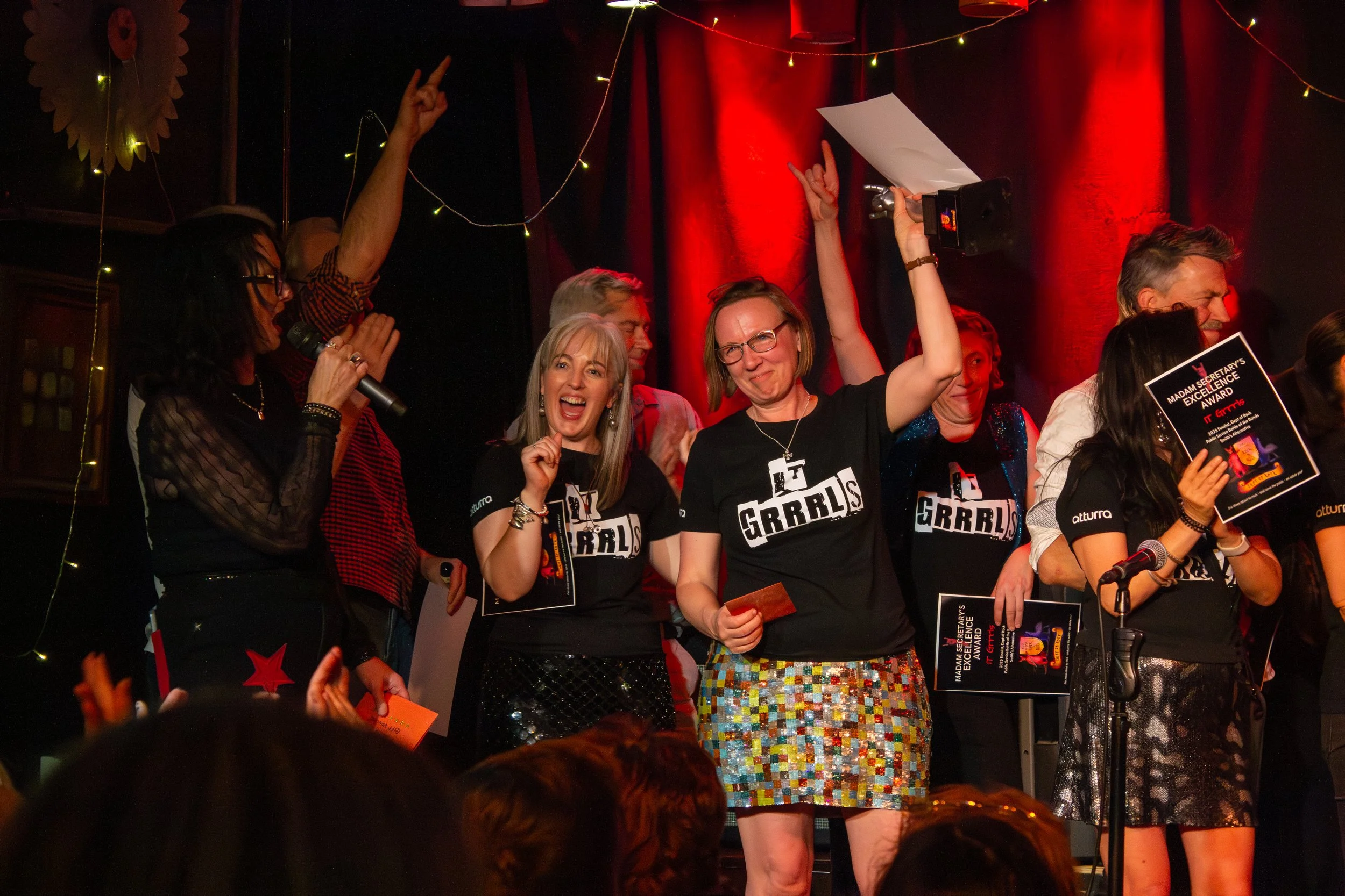 Group of women on stage celebrating at an event, some holding certificates, one speaking into a microphone, with red and black curtains, and fairy lights in the background.