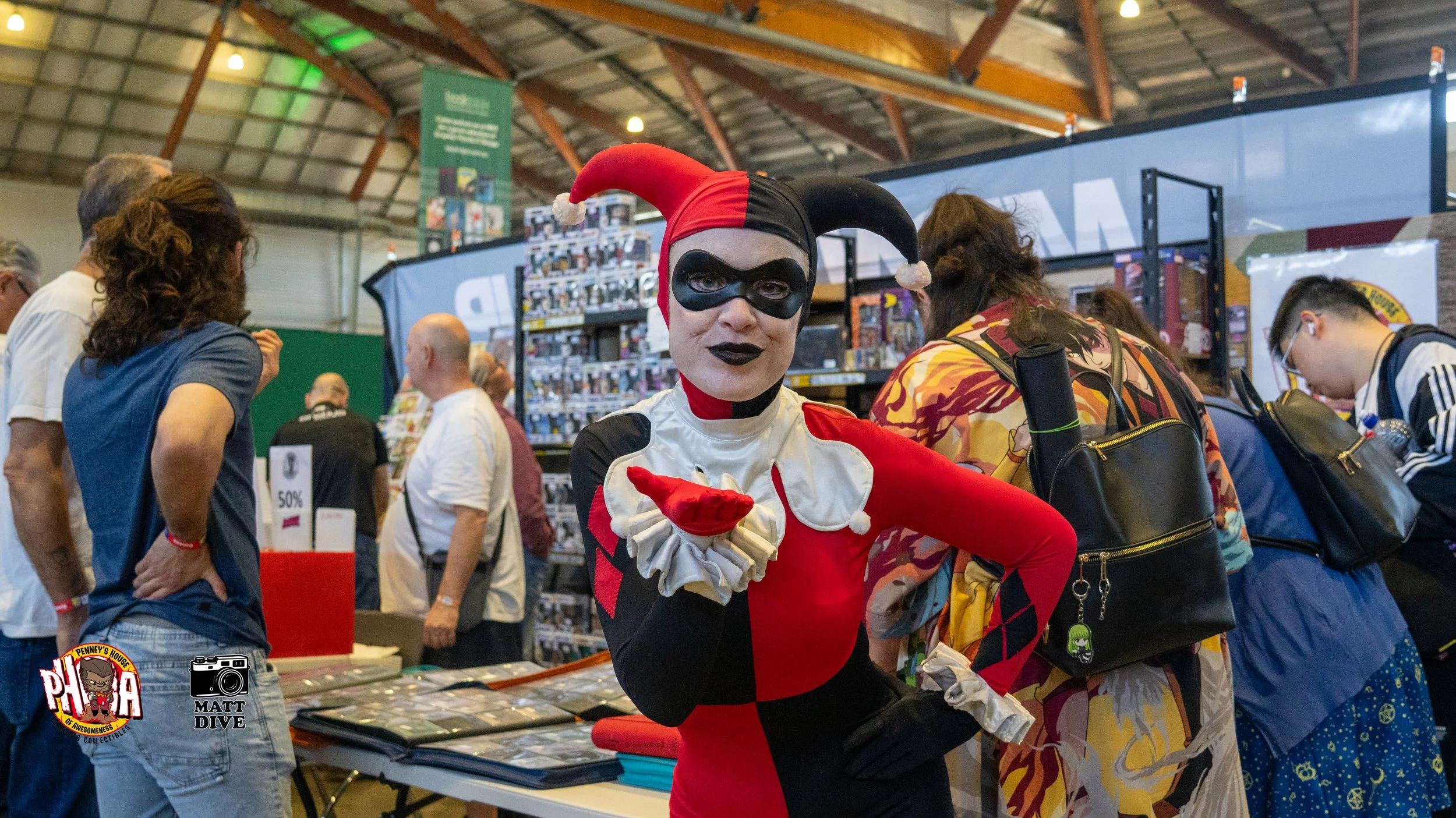 Person dressed as Harley Quinn in a red and black costume with a jester hat, posing with a playful expression at a convention or event, surrounded by other attendees and displayed merchandise.