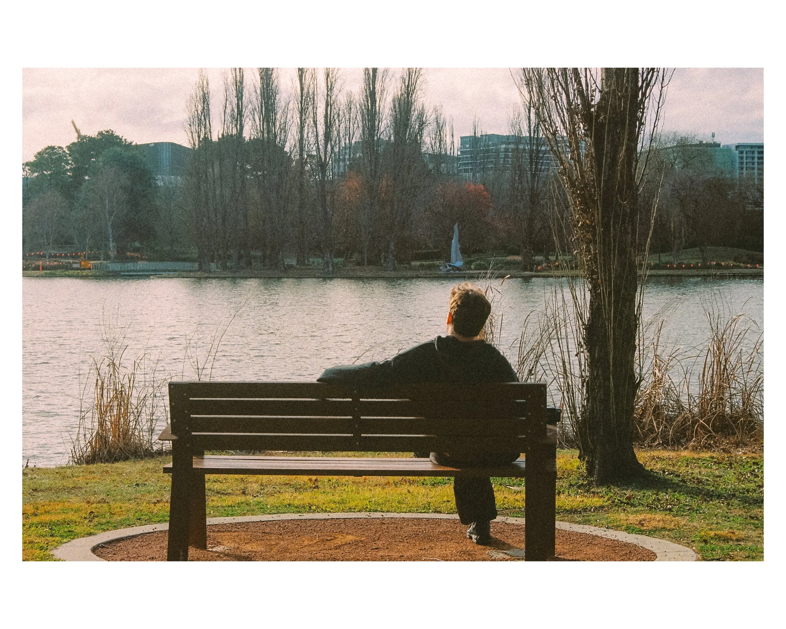 A person sitting on a park bench by a lake, looking out over the water and trees in the distance during dusk or early evening.