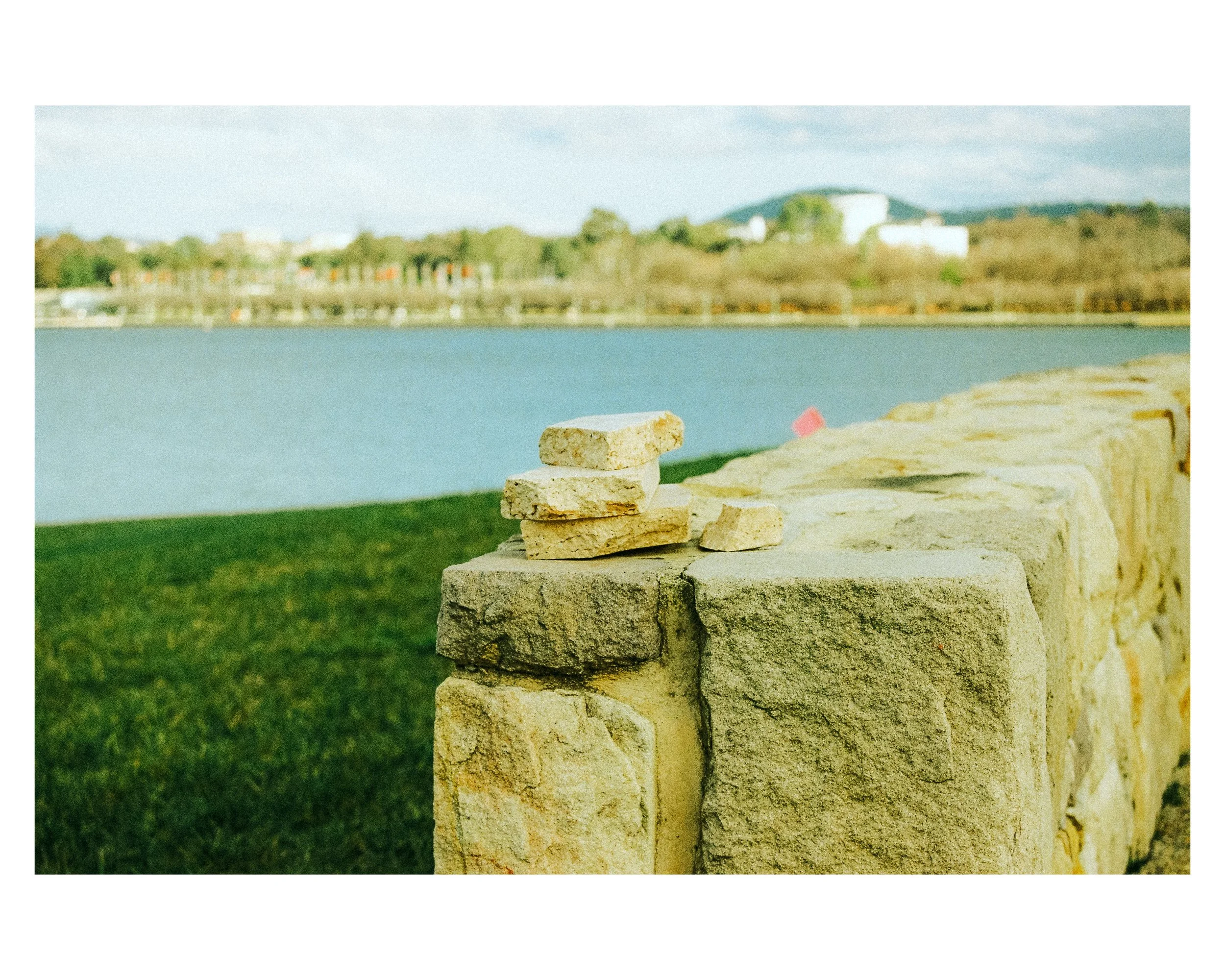 A stone wall with some flat stones stacked on top near a body of water, with greenery and buildings in the background.