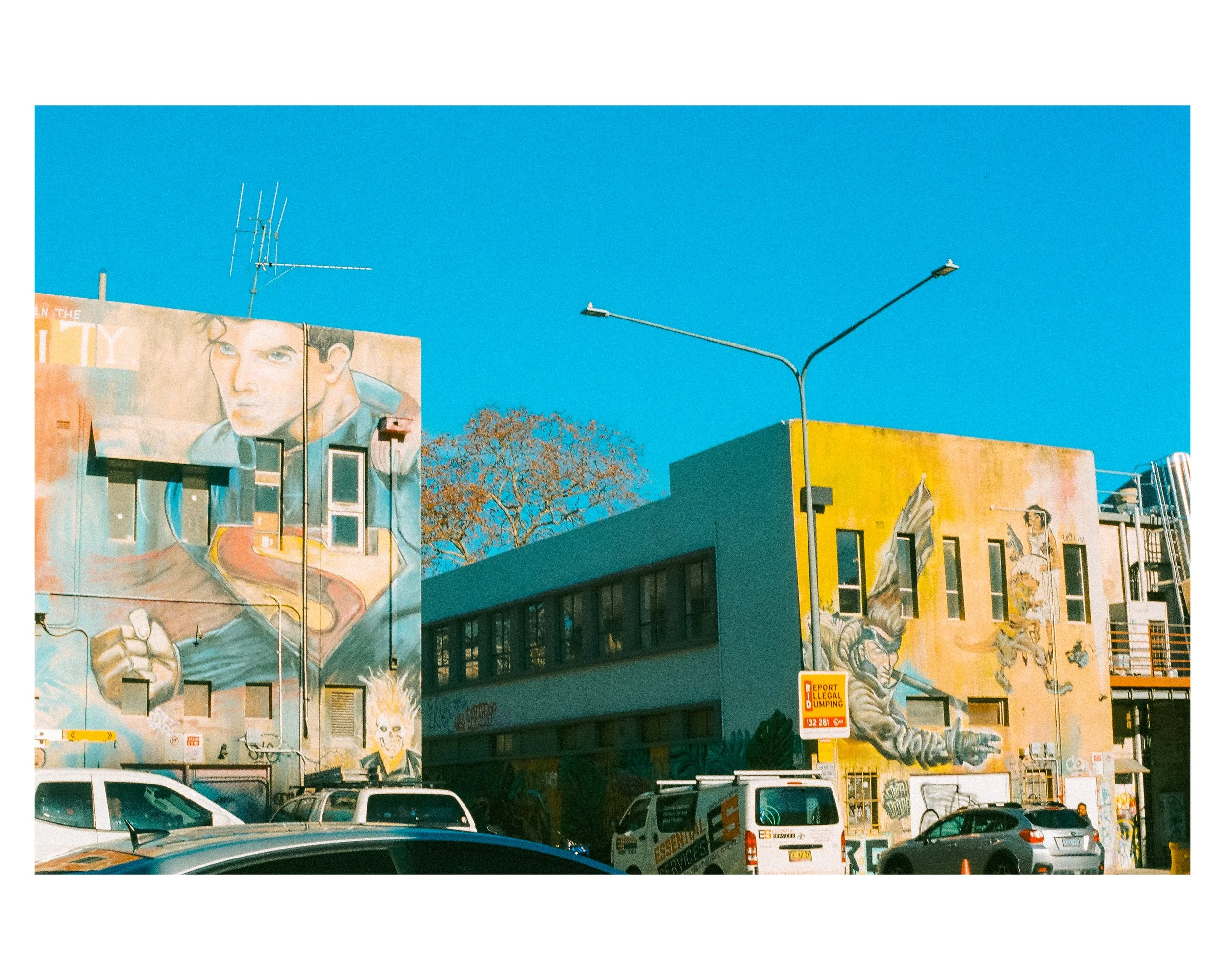 Colorful murals on buildings depicting a man holding a microphone and a person riding a horse, with parked cars below and a clear blue sky above.