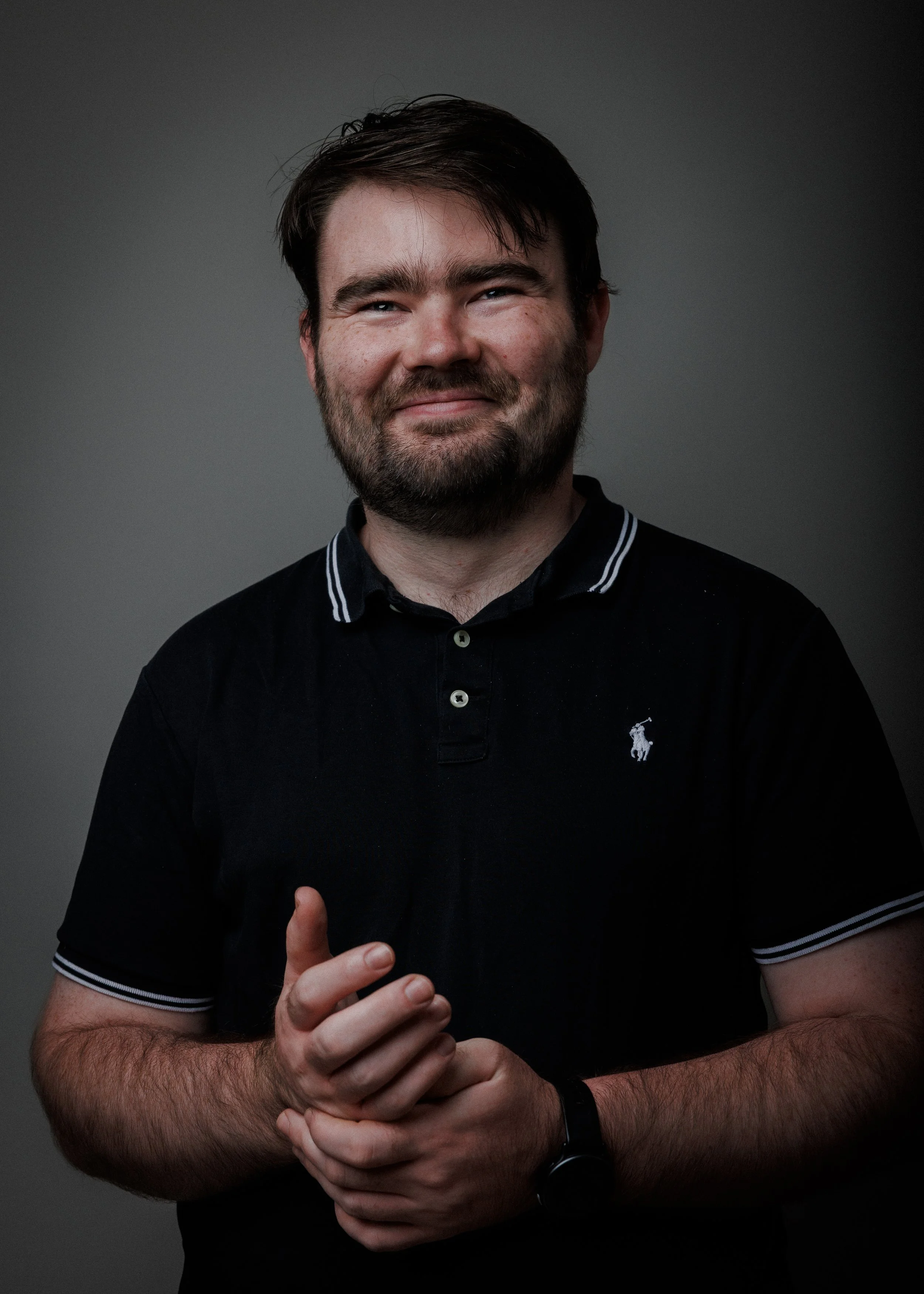 A man with dark hair and a beard, wearing a black polo shirt with white trim, smiling with hands clasped in front of him, against a plain gray background.