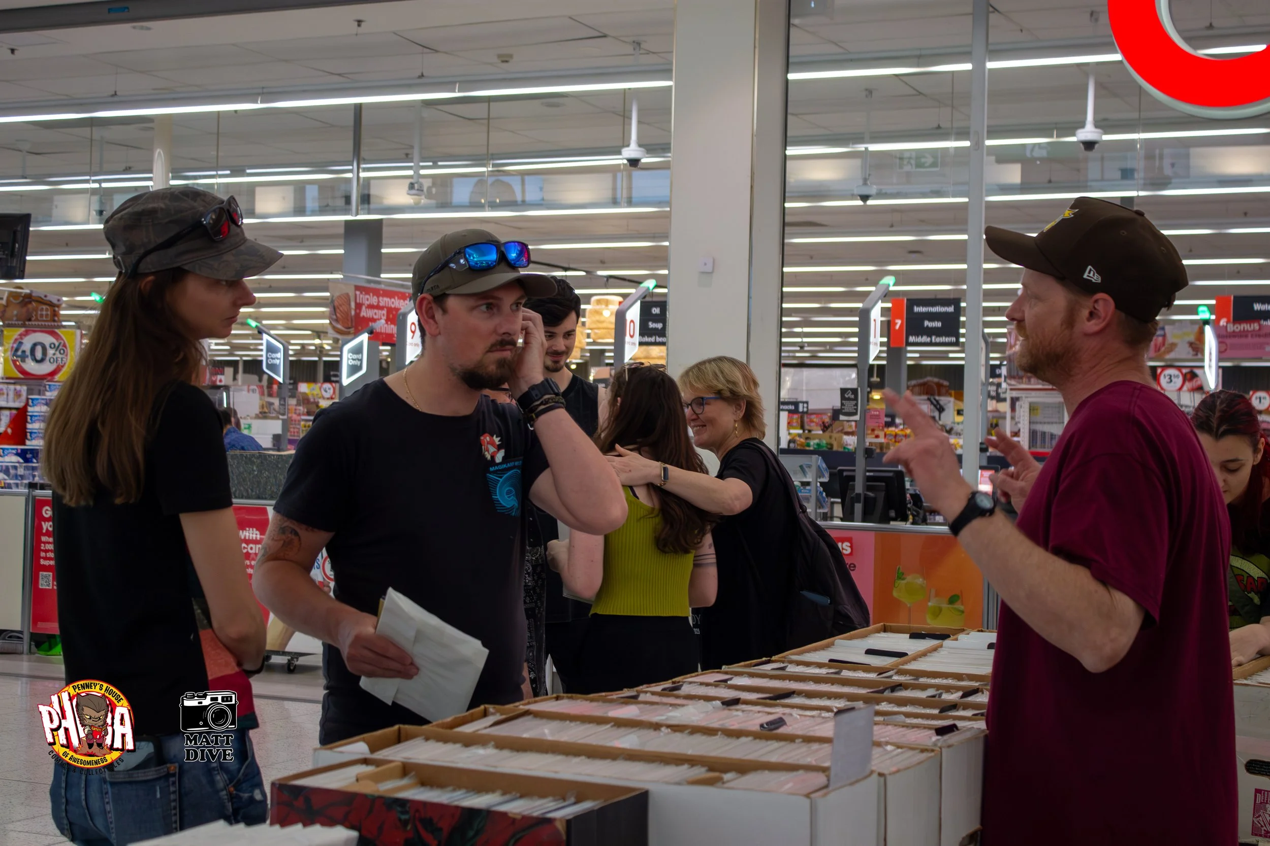 People shopping at an outdoor book sale in a supermarket aisle, with books displayed on tables.
