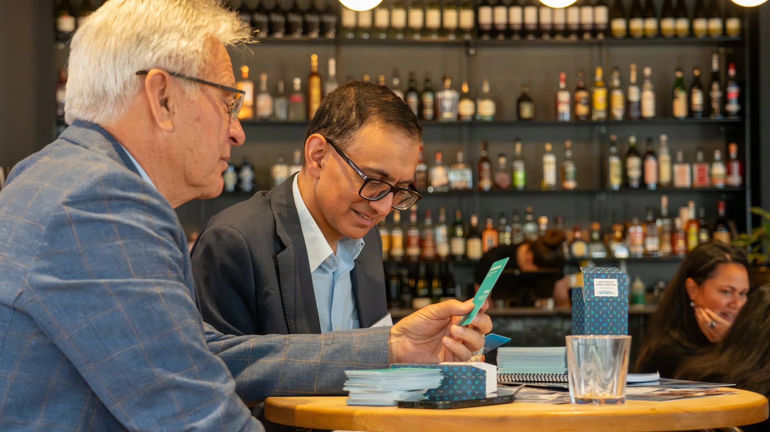 Two men sitting at a bar or restaurant table, looking at a card or piece of paper together, with a woman smiling in the background.