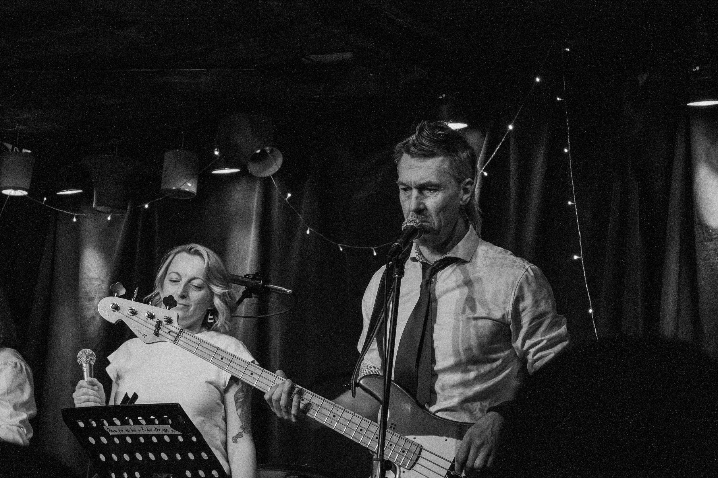 A black and white photo of a male guitarist and singer performing on stage with a female singer. The male is singing into a microphone, wearing a shirt and tie, with a focused expression. The female is holding a microphone, with her eyes closed and a