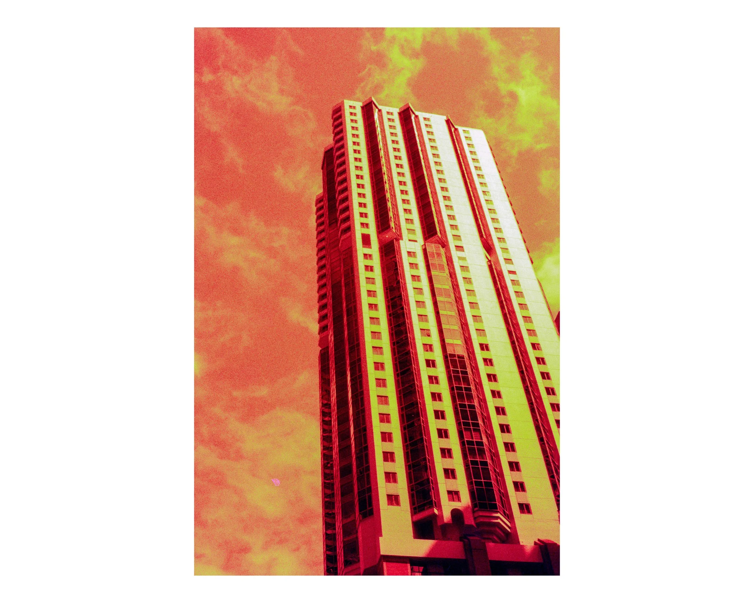 A tall, modern skyscraper with multiple windows, set against an orange-red sky with some clouds.