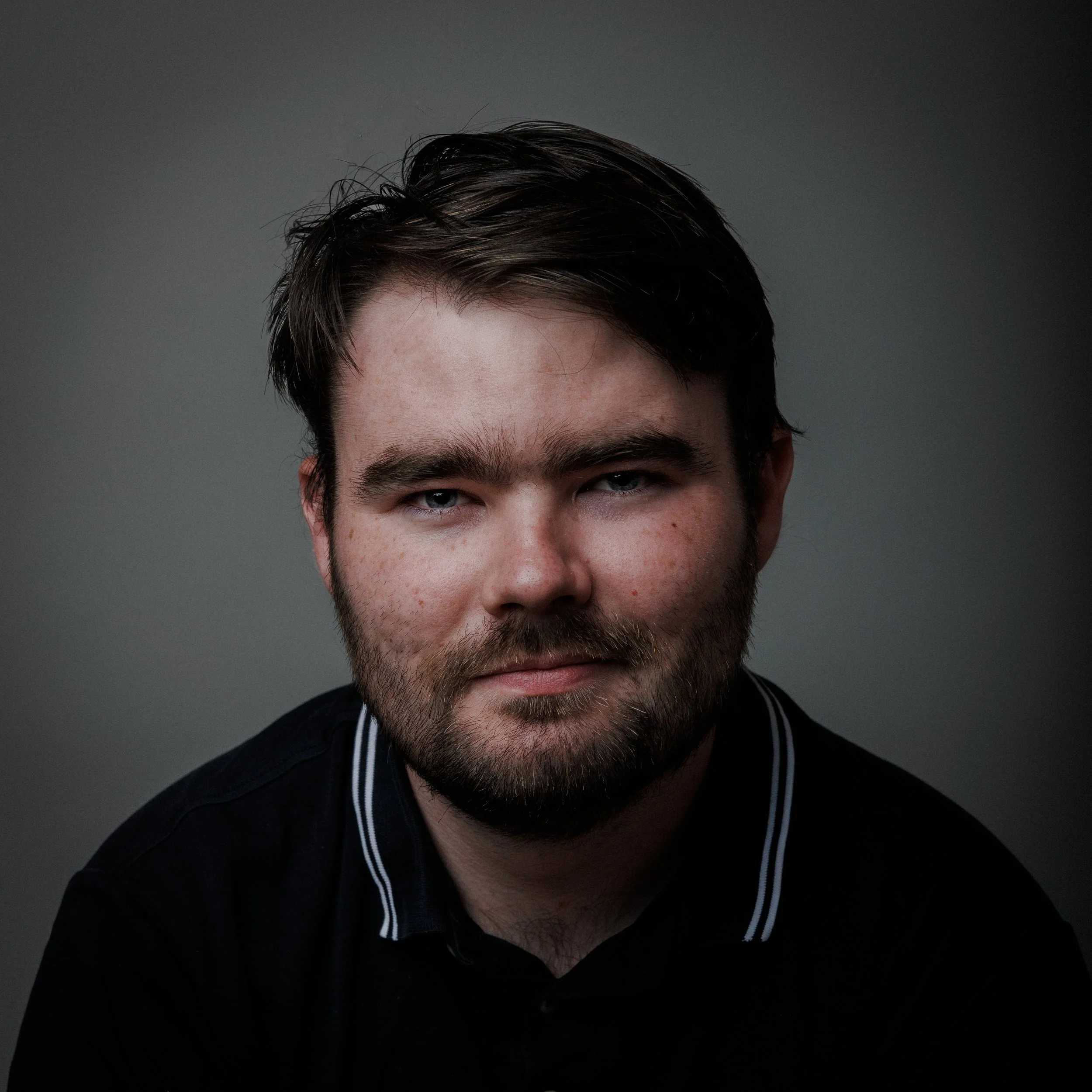 Close-up portrait of a young man with dark hair and beard, wearing a black shirt with white striped collar, against a dark gray background.