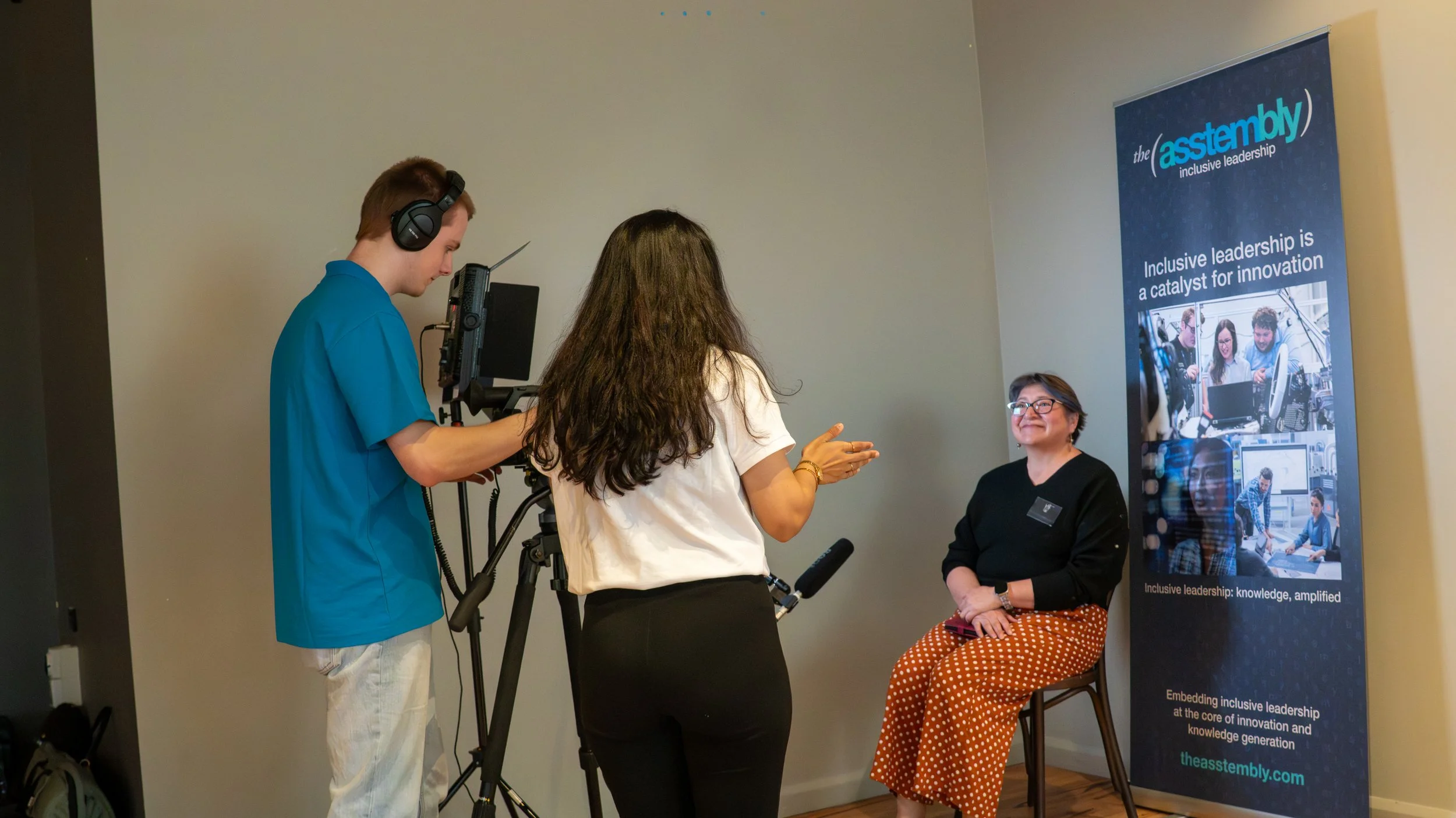 A woman being interviewed on camera in front of a blue banner that reads 'the Assembly' and discusses inclusive leadership. The woman sits on a chair, smiling, and is wearing glasses, a black top, and orange polka dot pants. The banner features image