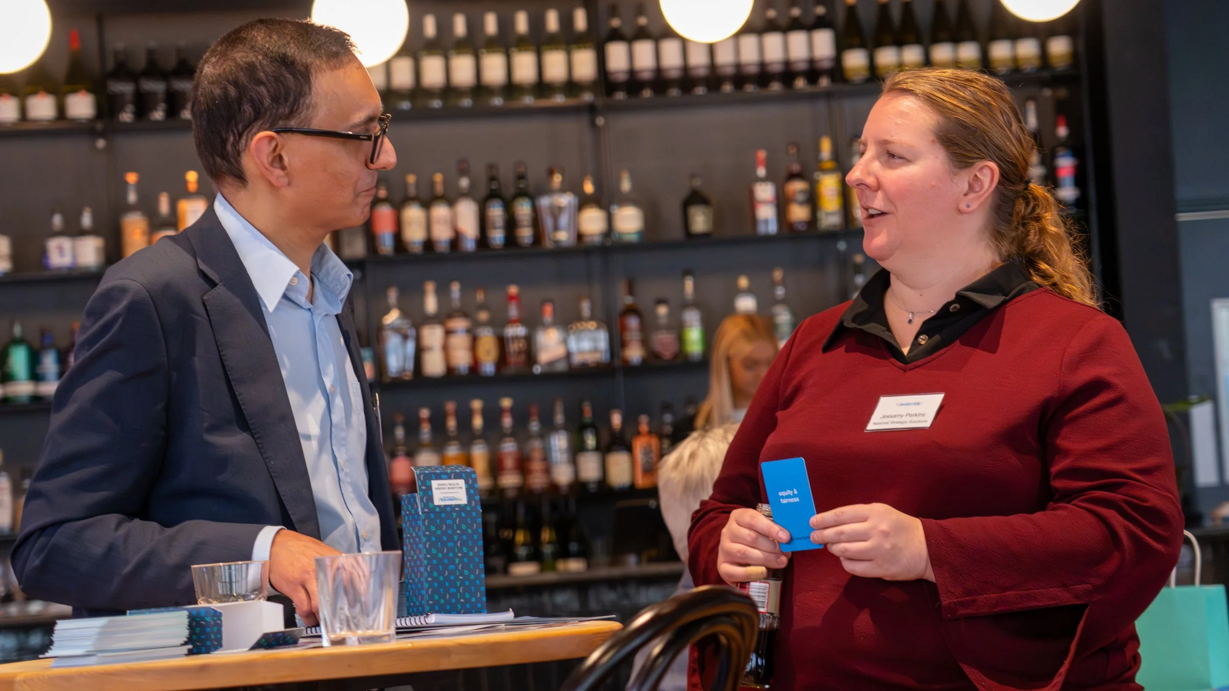 A man and a woman are talking at a bar or restaurant. The man is wearing glasses, a dark blazer, and a light blue shirt. The woman has a name tag that reads "Lacey P...