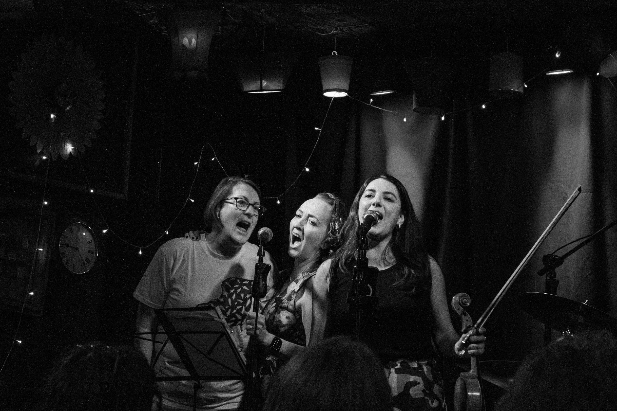 Three women singing passionately on a stage with microphones, a violin, and music stands in a dimly lit venue decorated with hanging lamps and string lights.