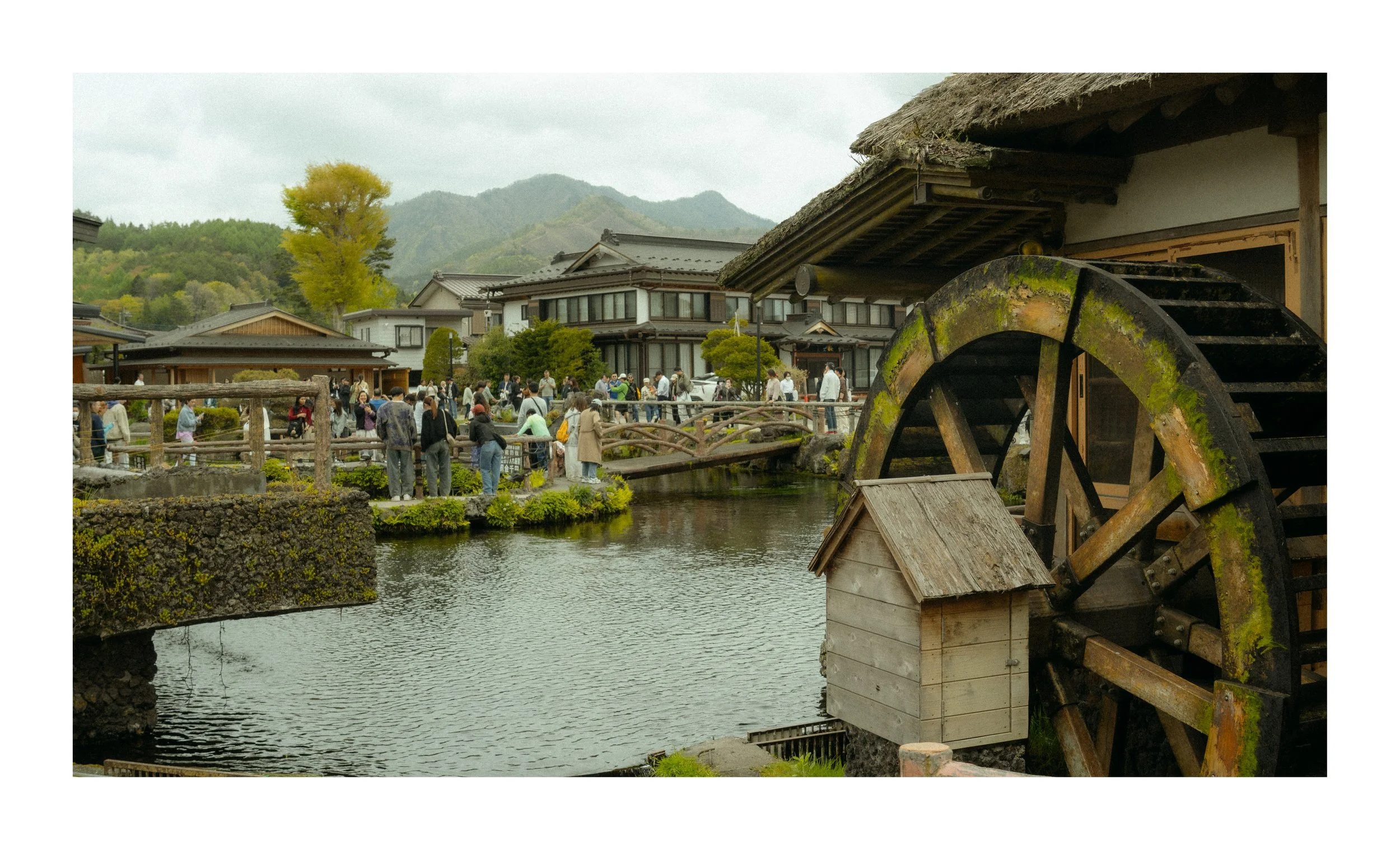 View of a traditional Japanese village with a pond, a group of people on a bridge, lush greenery, and mountains in the background. An old wooden water wheel is in the foreground.