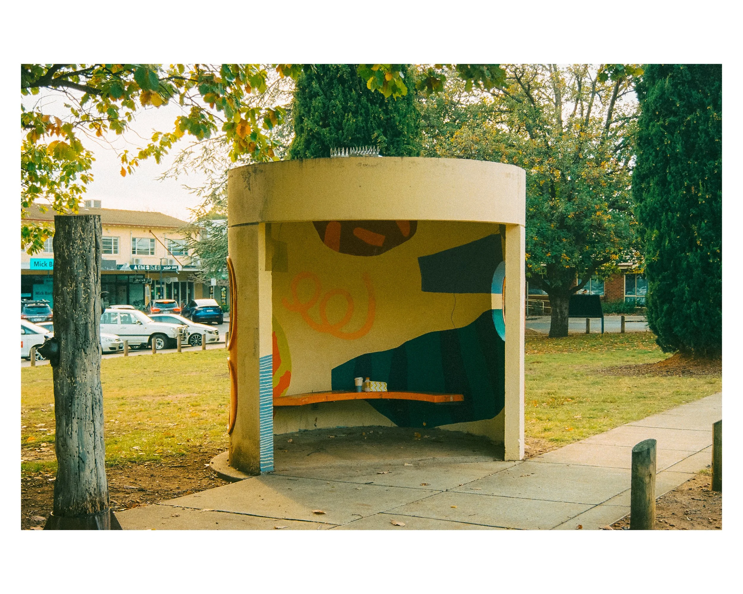 Empty bus shelter with colorful mural, trees, parked cars, and buildings in the background.