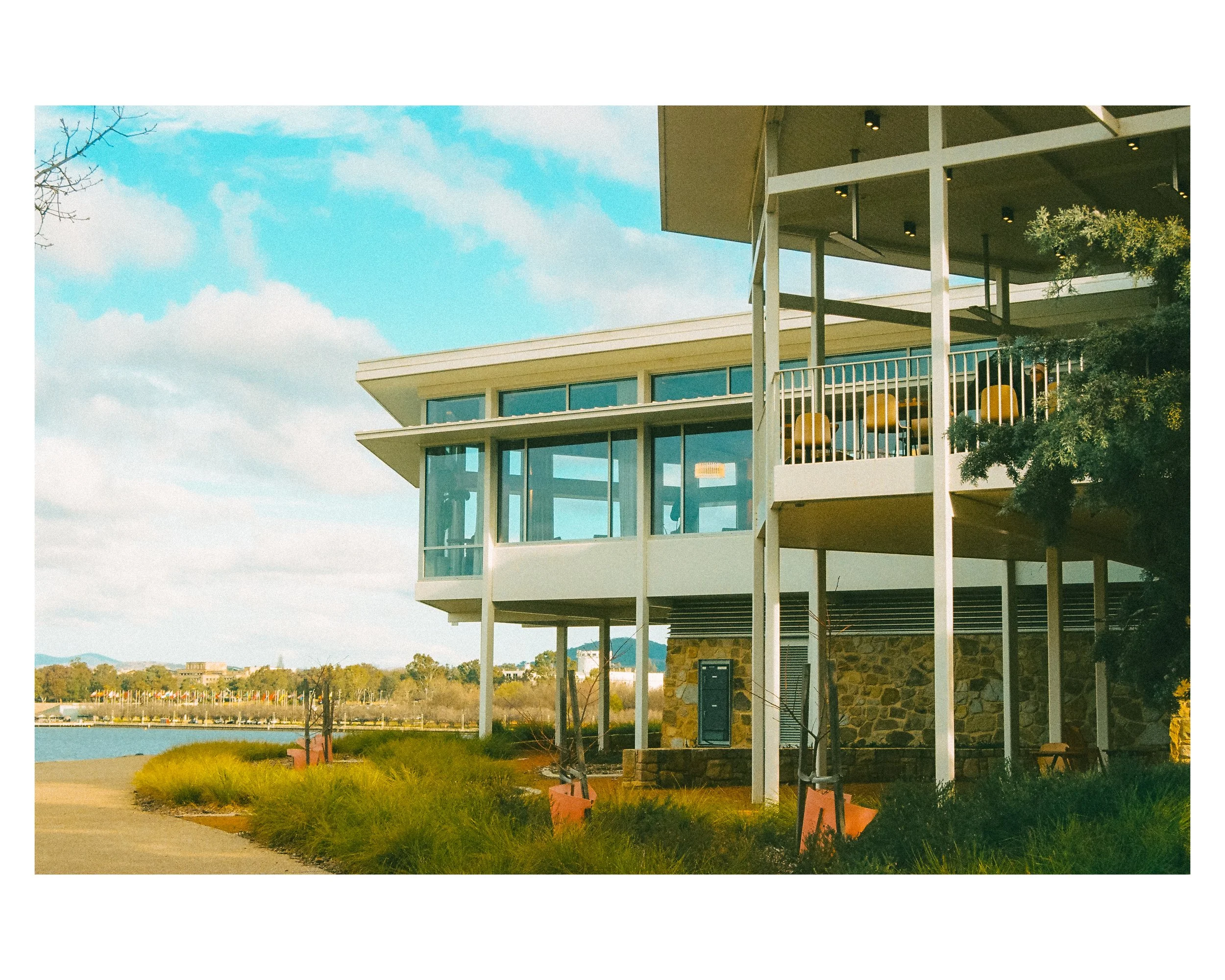 Modern waterfront building with large glass windows, located by a river, with a walking path and grassy area in foreground.
