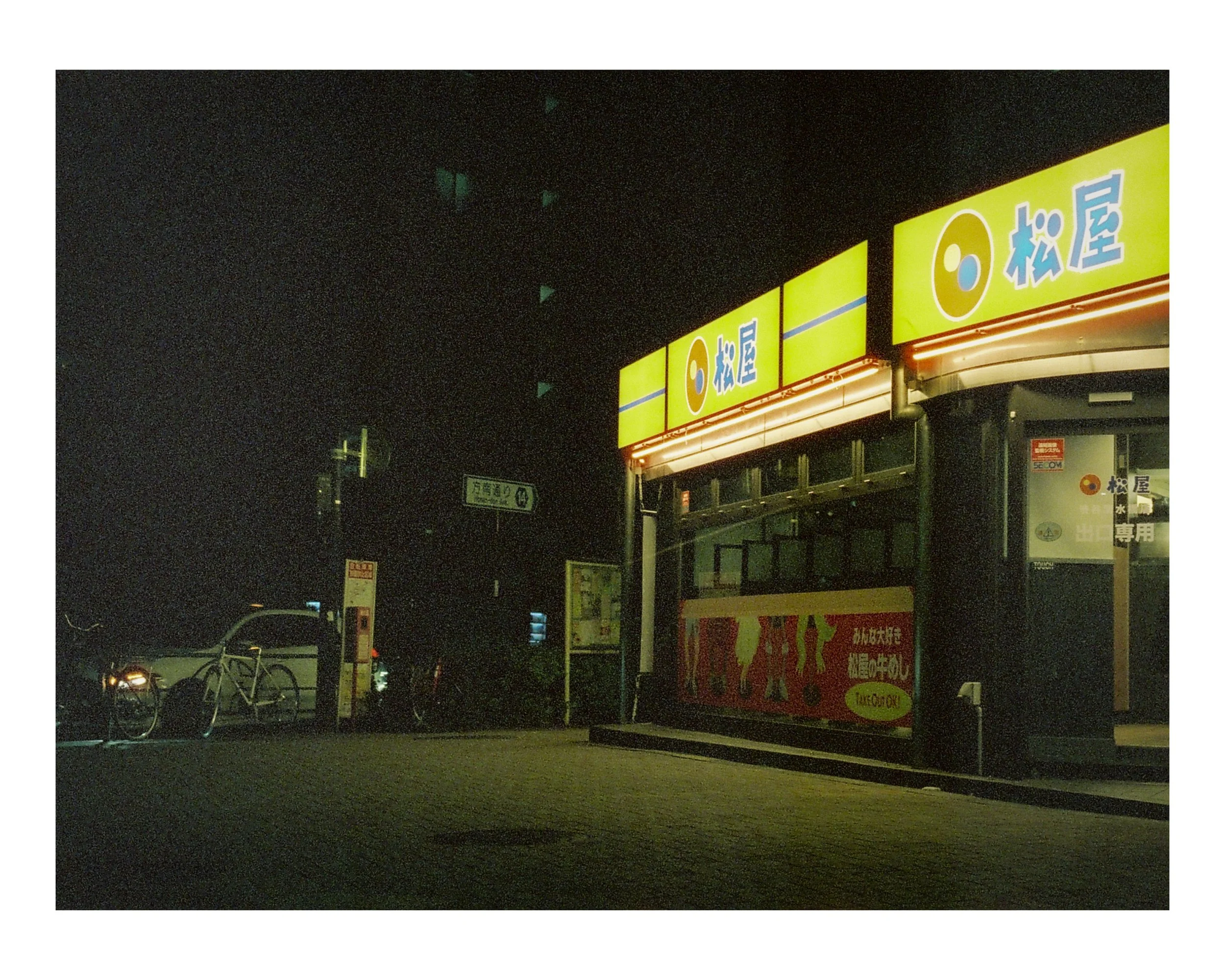 Nighttime scene of a Takoyaki stand with bright yellow signage and Japanese text, parked bikes, and a few cars in the background.