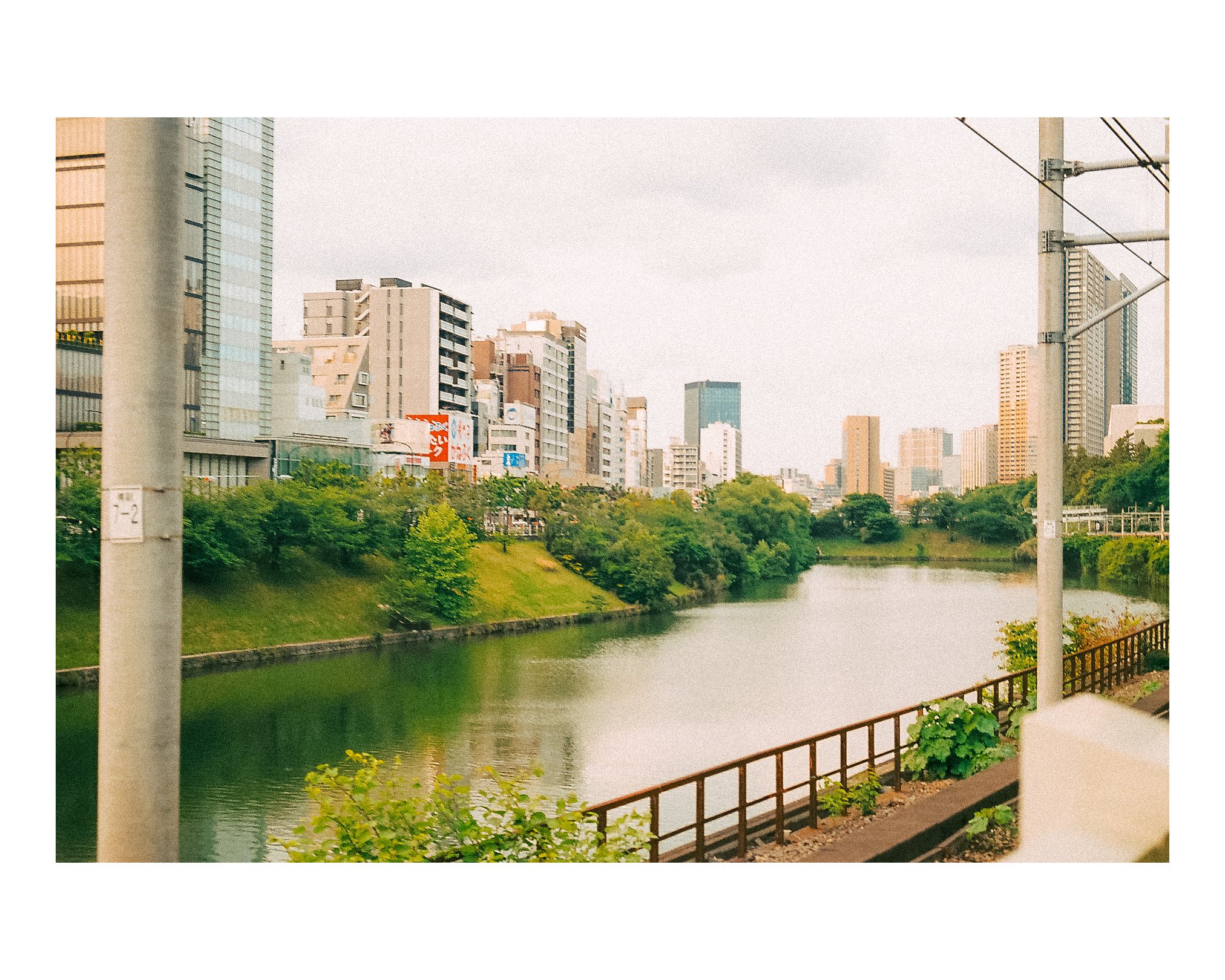 Urban cityscape with a river, modern high-rise buildings, green trees, and a cloudy sky.