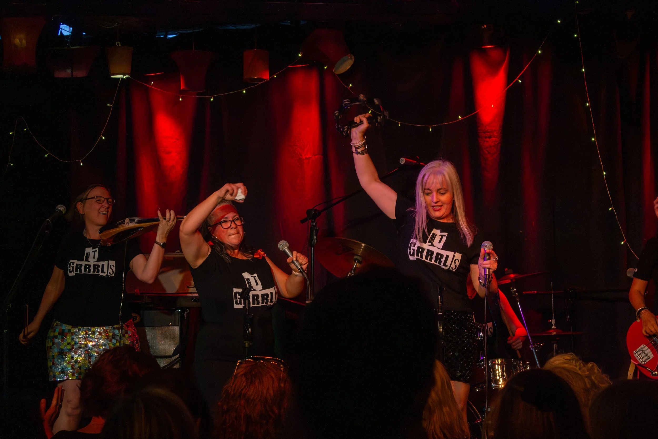Women on stage at a concert, holding microphones and raising a tambourine, with red and black curtains and string lights in the background, wearing matching black shirts with a logo.